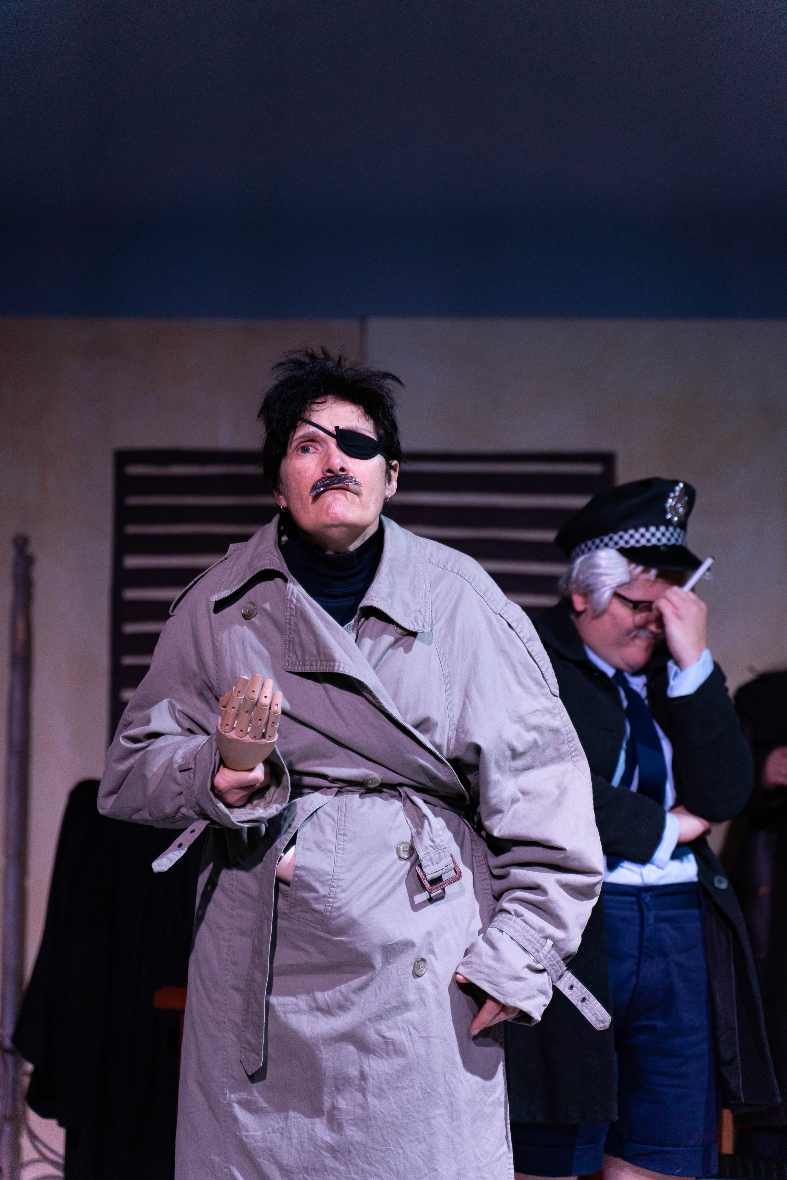 A theatrical scene with a person dressed as a stereotypical detective wearing a trench coat, eyepatch, and fake mustache, holding a robotic hand, and a woman dressed as a police officer with glasses, a police hat, and a cigarette, standing in front o