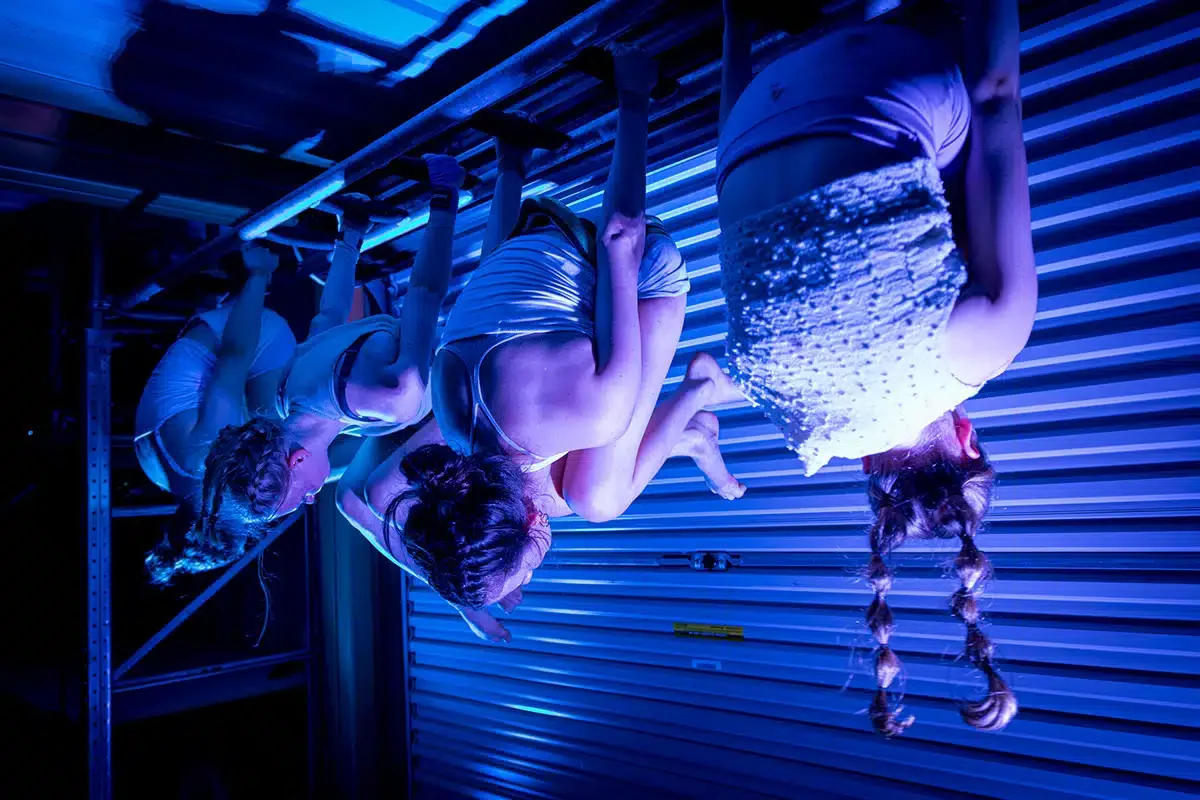 Four women hanging upside down from a horizontal pole, illuminated by blue lighting, with their hair braided and dressed in white athletic wear.