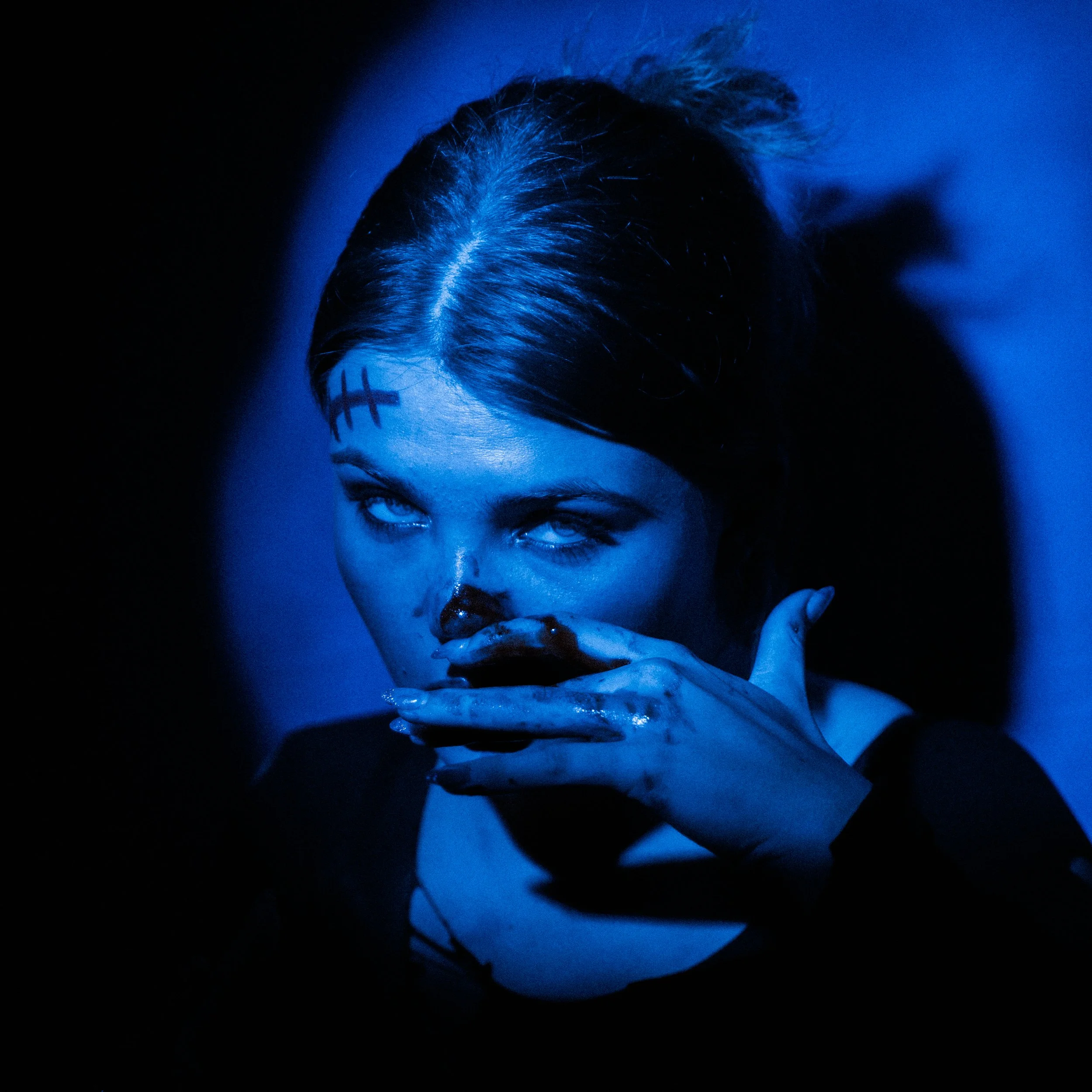 A woman with dark hair and blue face makeup, with black markings on her forehead, covers her mouth with her hand, looking upward with a blue-tinted lighting.