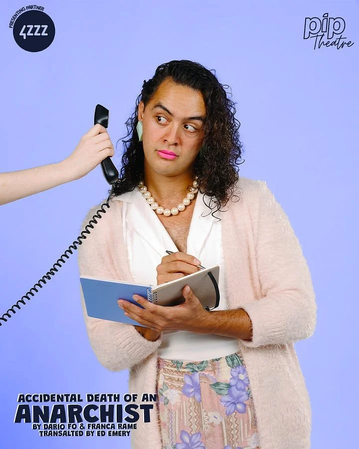 A woman with dark, curly hair looks confused or skeptical as she holds a notepad and pen. Someone off-camera is holding a telephone receiver to her ear. She is wearing a white blouse, a pearl necklace, and a light pink cardigan. The background is lig