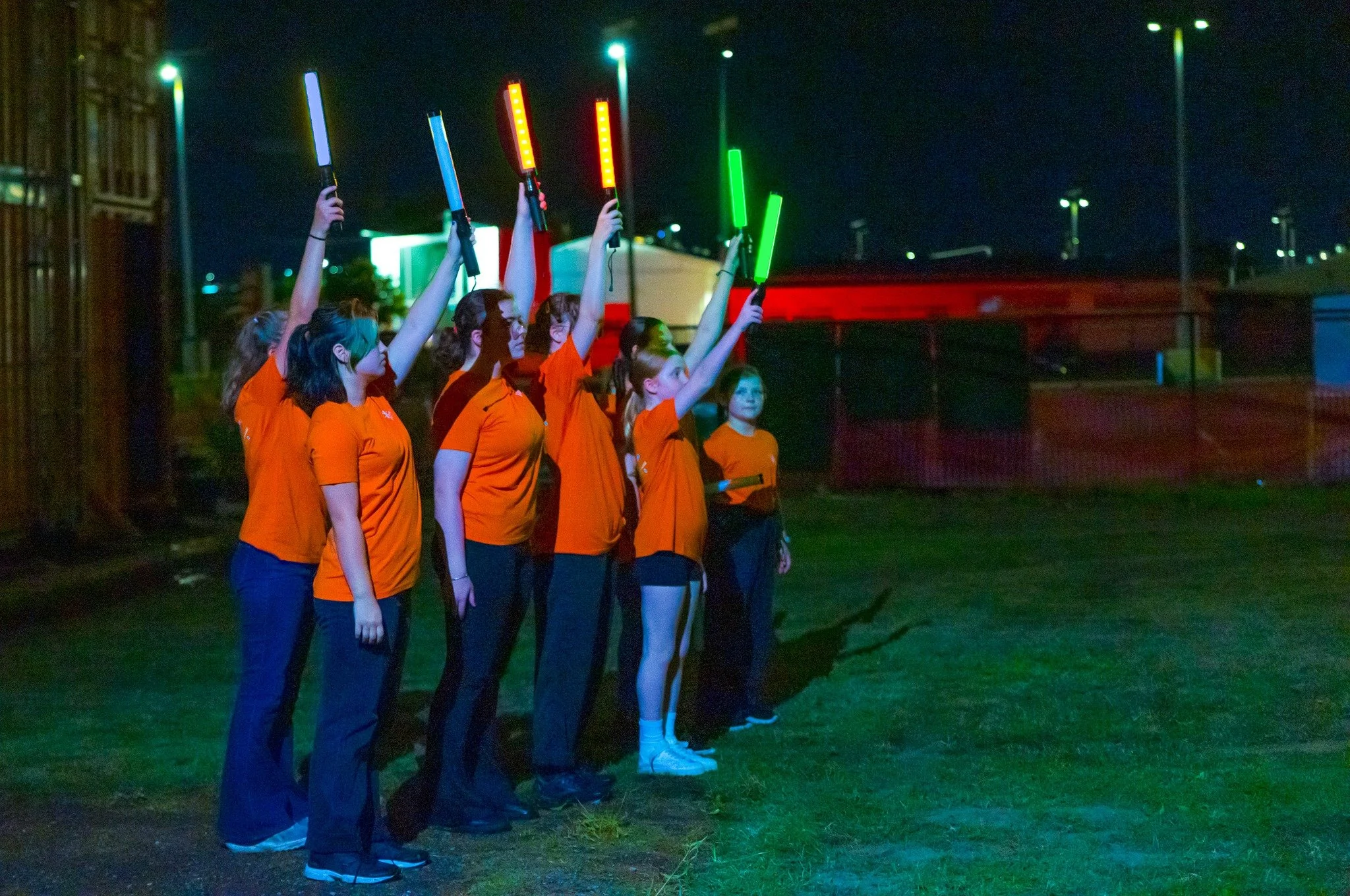 Group of women wearing orange shirts holding illuminated light sticks during a night event outdoors.