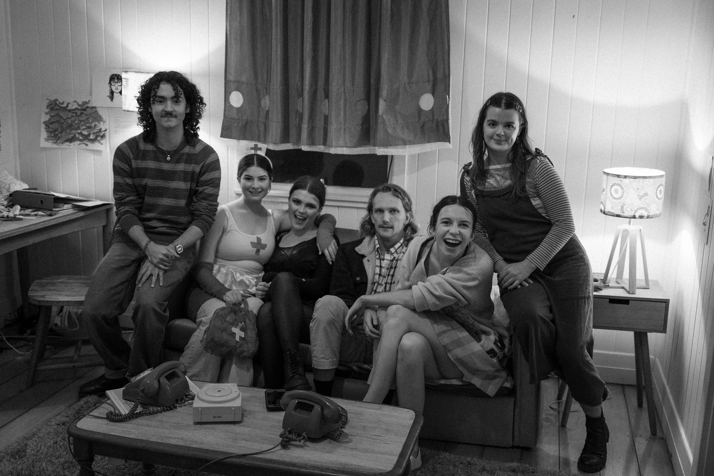 Group of six people posing together in a room, some dressed as nurses, with a wooden wall, window with curtain, and various furniture including a table with vintage phones and a lamp.