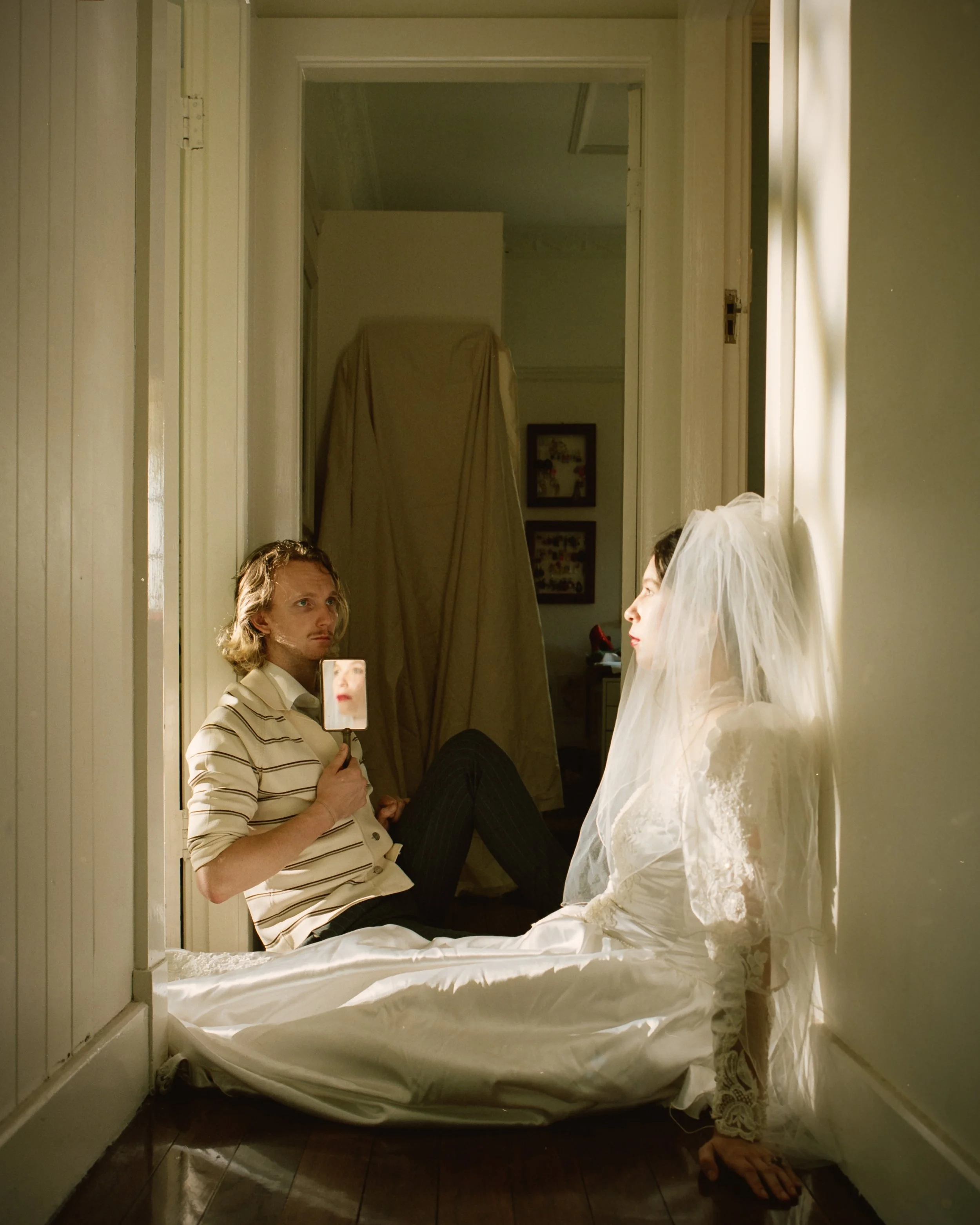 A woman in a wedding dress sitting on the floor with her back against the wall, wearing a veil, looking at a man in casual clothing holding a mirror, both appear to be in a room with peeling paint and dim lighting, with a makeup-covered face reflecte
