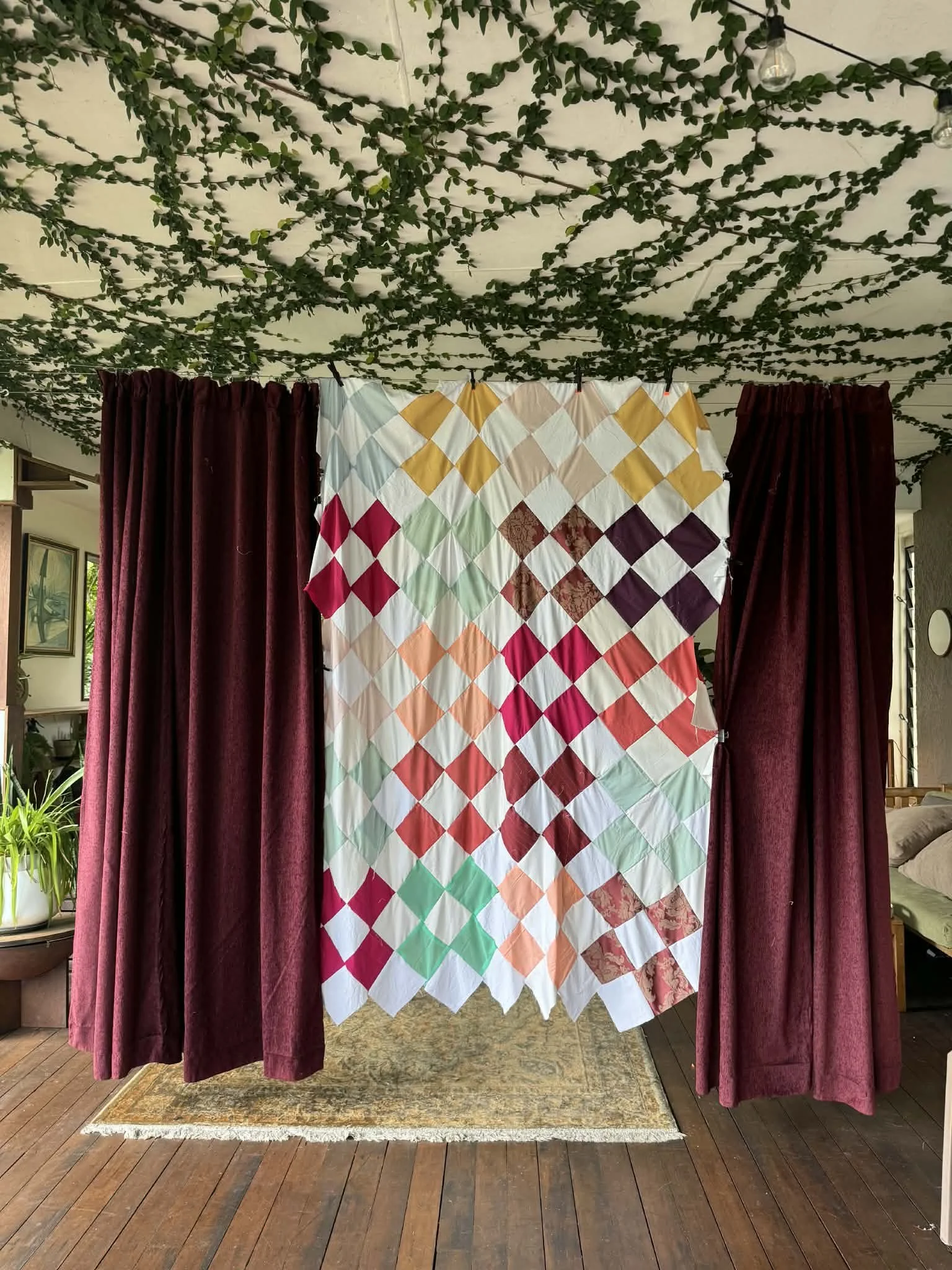A quilt with a diamond pattern of multiple colors hanging between two burgundy curtains in a room decorated with string lights and greenery on the ceiling.