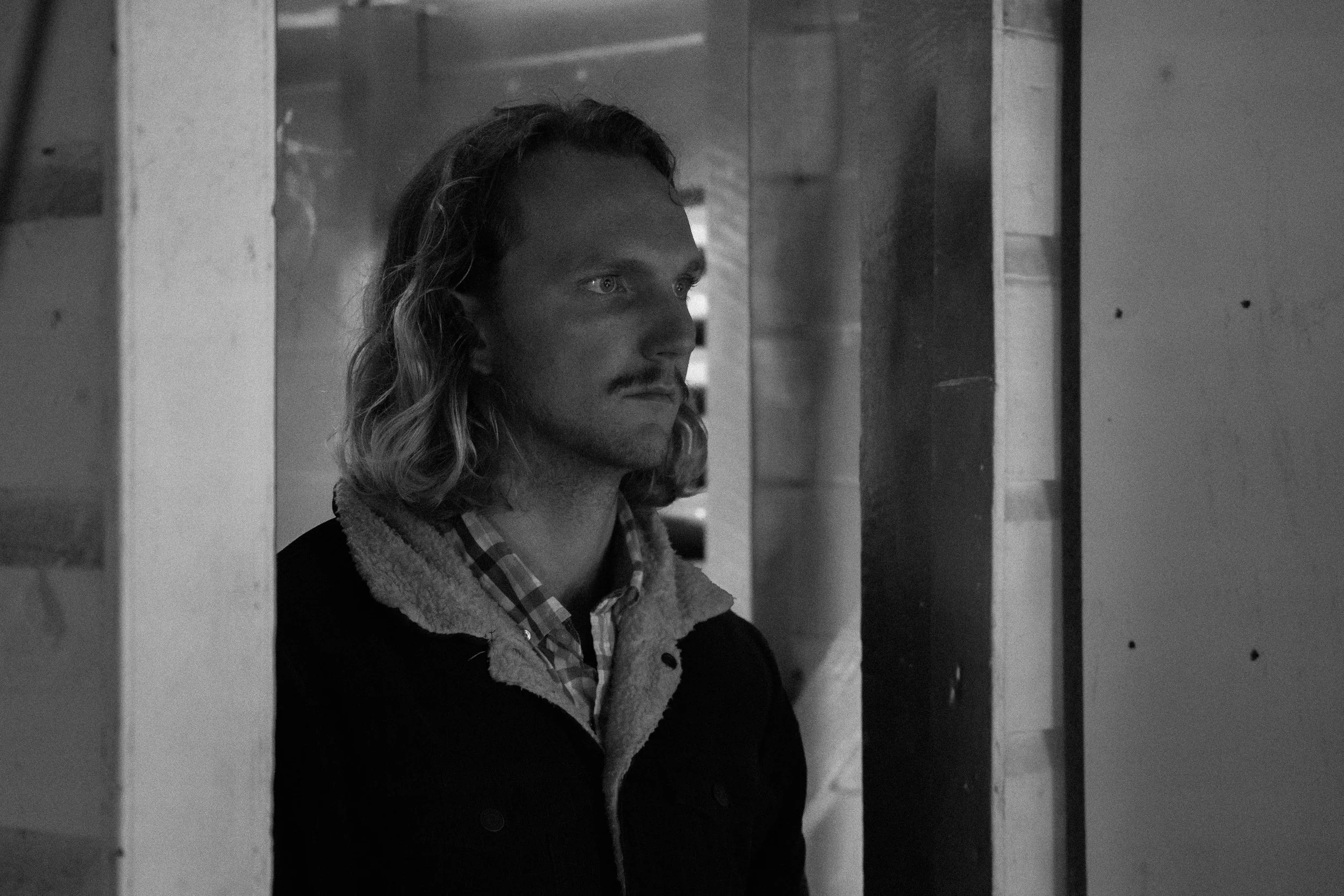 A man with long hair and a mustache, wearing a plaid shirt and a jacket with a fleece collar, standing in a doorway in a black-and-white photo.