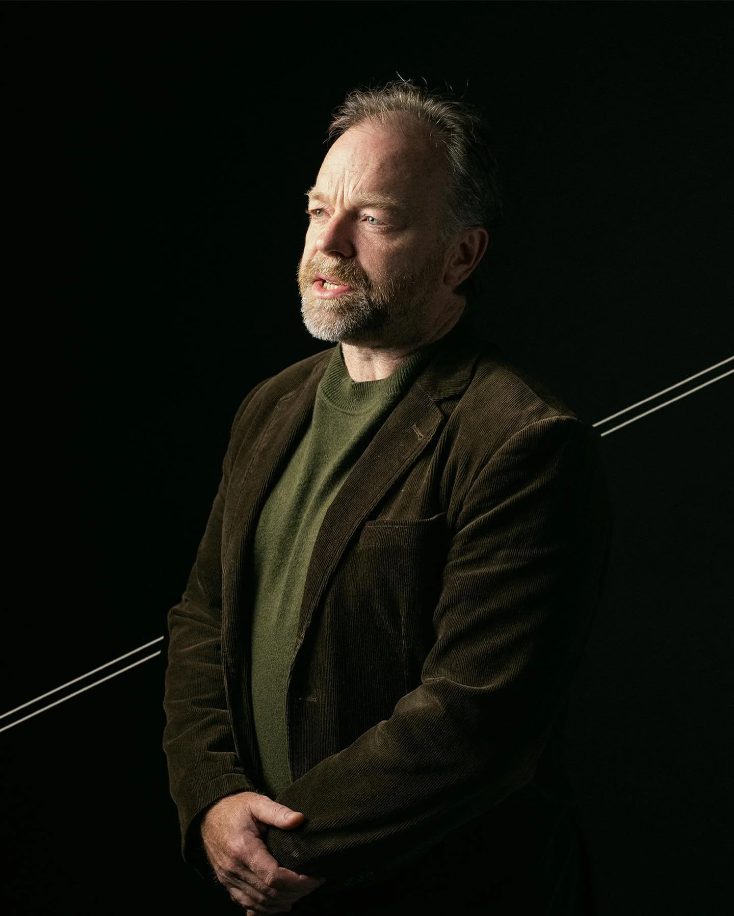 A middle-aged man with a beard and mustache, wearing a brown blazer and green sweater, standing against a black background with silver lines, looking to the side.