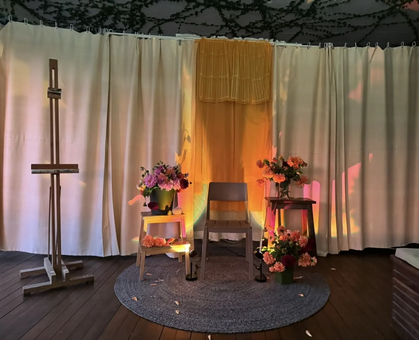 Stage with a beige curtain backdrop, three flower arrangements in pink, purple, and white flowers on small tables and the floor, a gray rug, and a wooden chair in the center, decorated with warm lighting.