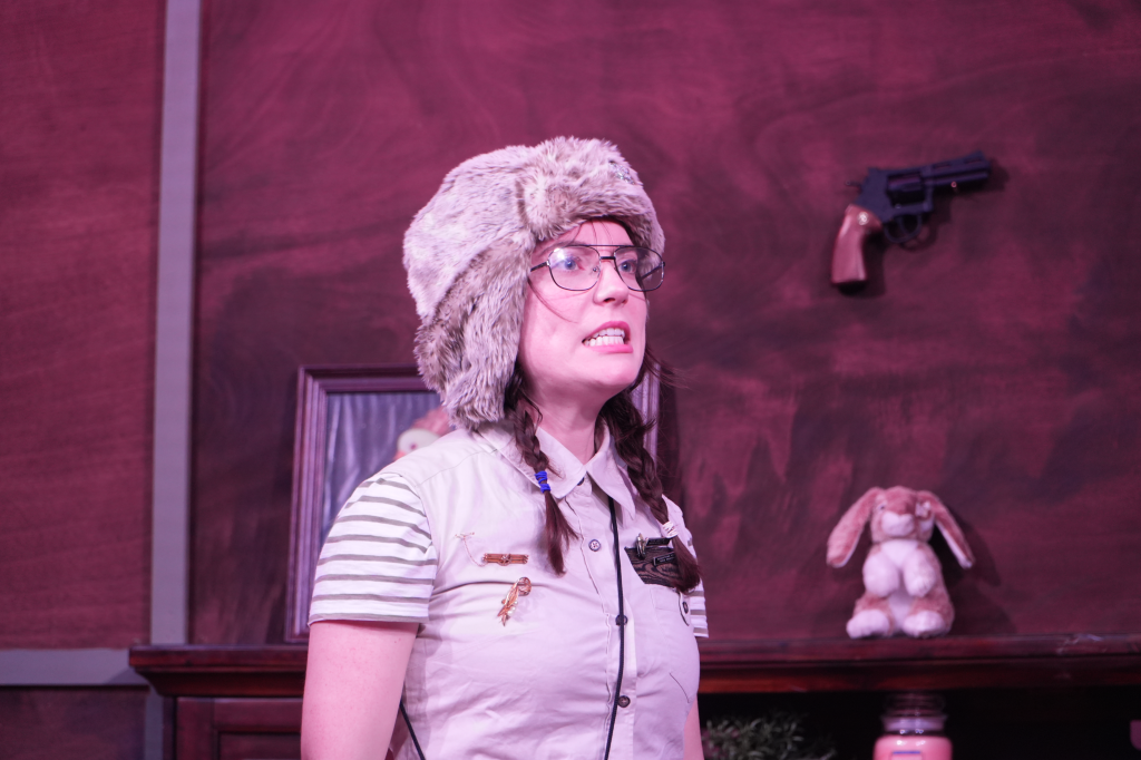 A woman with glasses, braided hair, wearing a furry hat and a beige shirt with striped sleeves, making a surprised or anxious facial expression, standing in front of a wooden wall with a toy gun and stuffed bunny on a shelf.