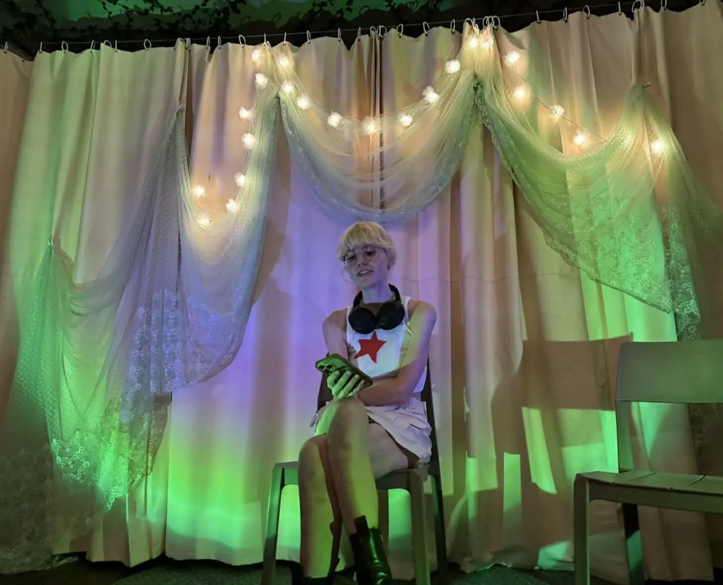 A person with blonde hair, glasses, wearing a white dress with a red star, headphones around their neck, sitting on a stool in front of a backdrop with curtains and string lights, holding a smartphone. The scene has colorful lighting, mainly green an