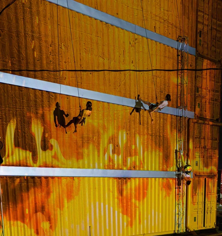 Six people are climbing on a steel structure with a large illuminated screen or projection of a fiery, lava-like background behind them.
