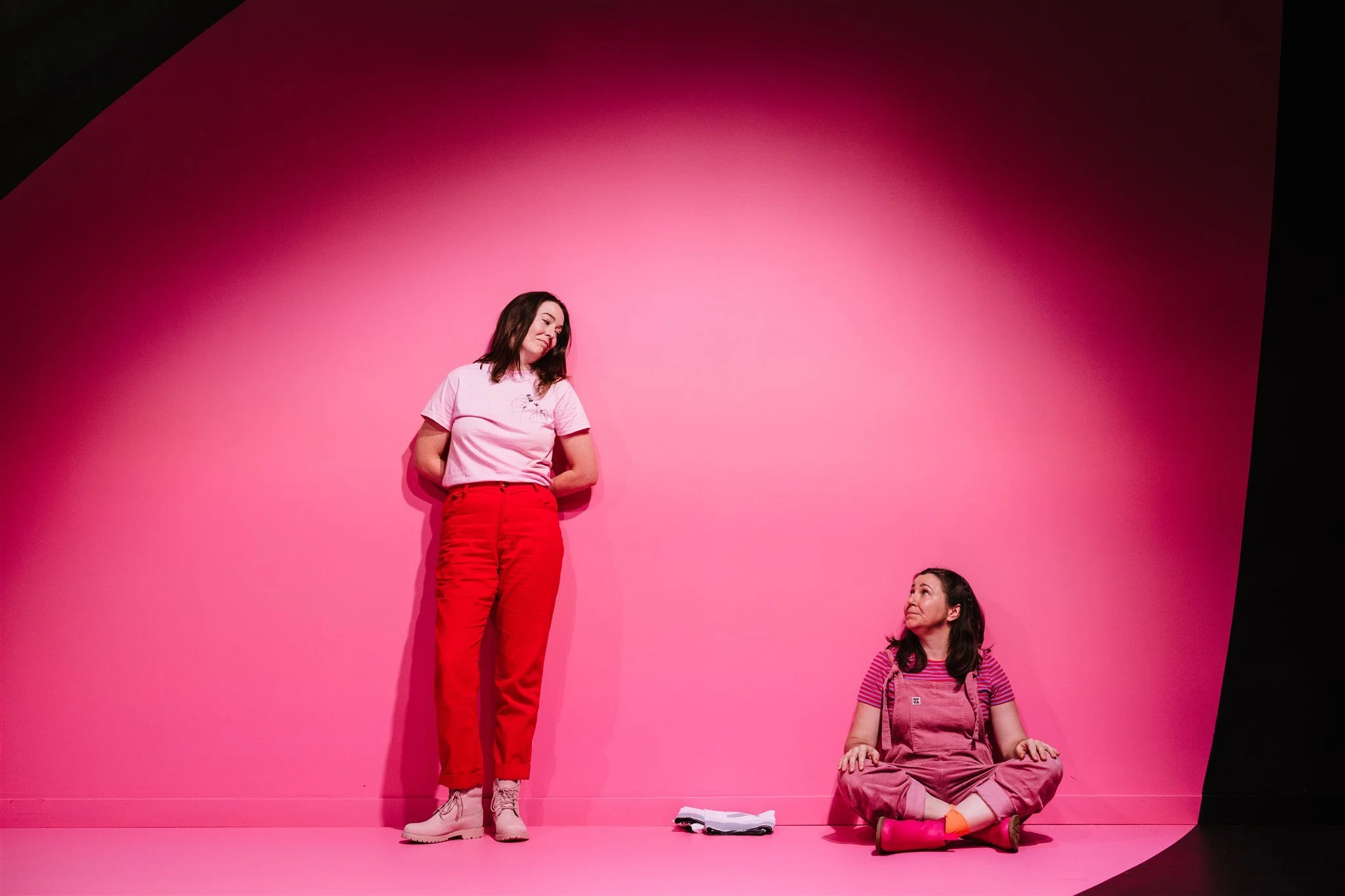Two women in pink outfits against a pink and black backdrop. One is standing, leaning slightly to the side with her hands behind her back. The other is sitting cross-legged on the floor, looking up at the standing woman. There are books on the floor 