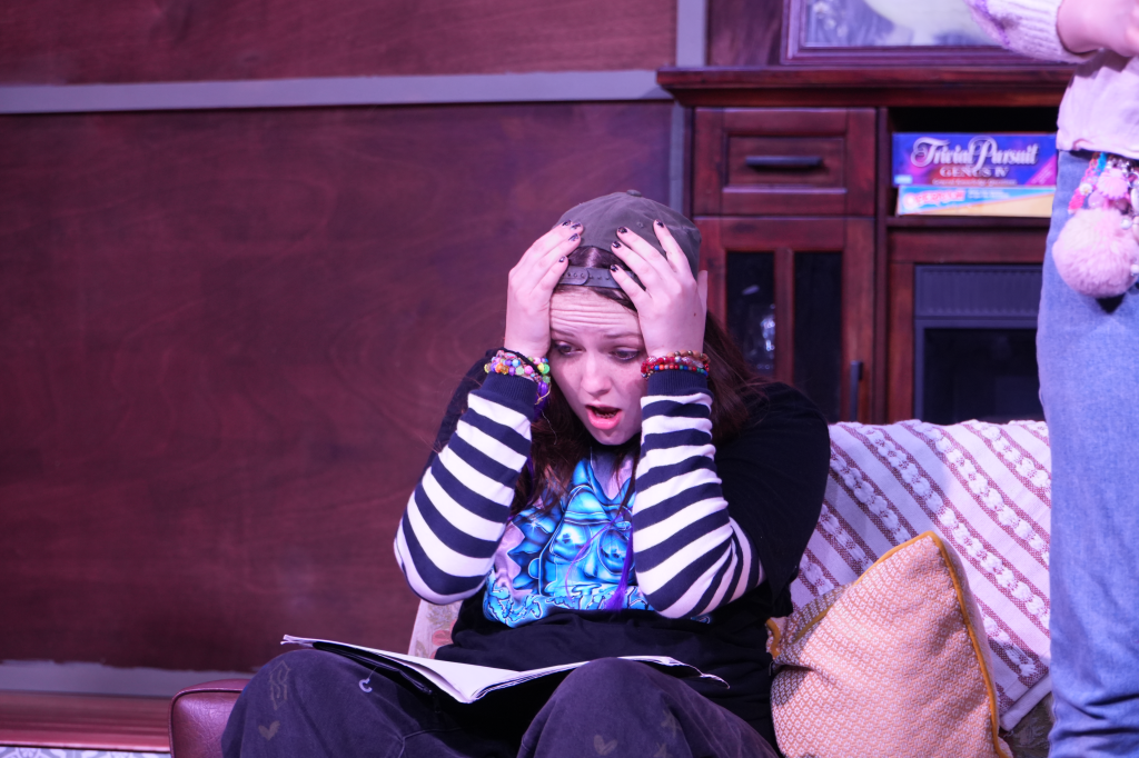A young woman sitting on a couch, looking shocked and distressed, with her hands on her head. She has dark hair and is wearing a gray baseball cap, a black shirt with striped sleeves, and colorful bracelets. There is an open book on her lap and a pat