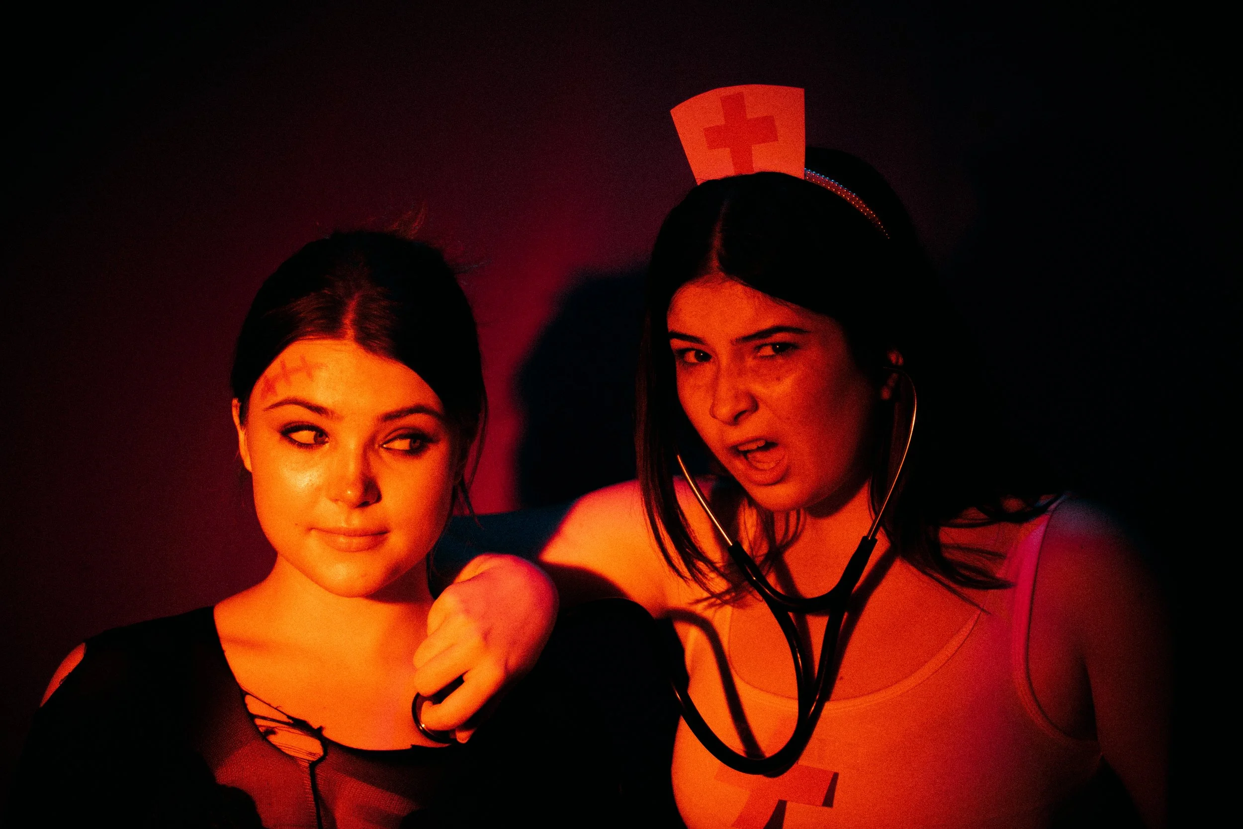 Two women in costume, one dressed as a nurse with a red cross hat and stethoscope, the other with makeup and tattoos, against a dark red background.