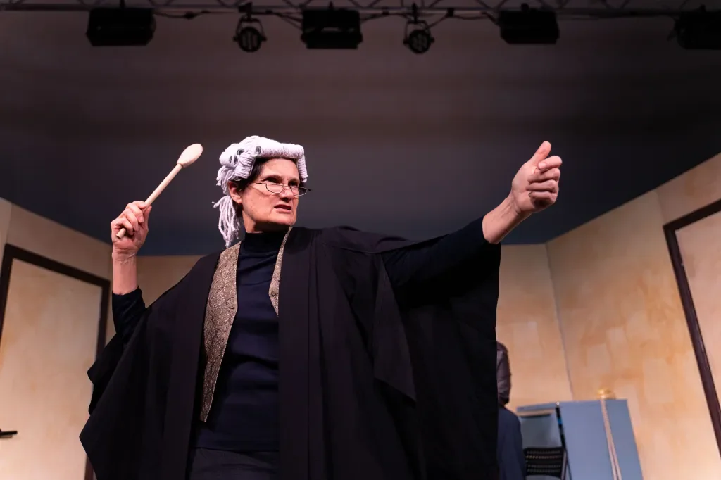 A person dressed as a judge or lawyer in a courtroom, wearing a wig, glasses, and black robes, holding a gavel in one hand and pointing with the other.