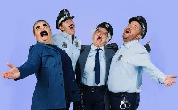 Four men dressed as police officers and police deputies, smiling, with arms outstretched, against a blue background.