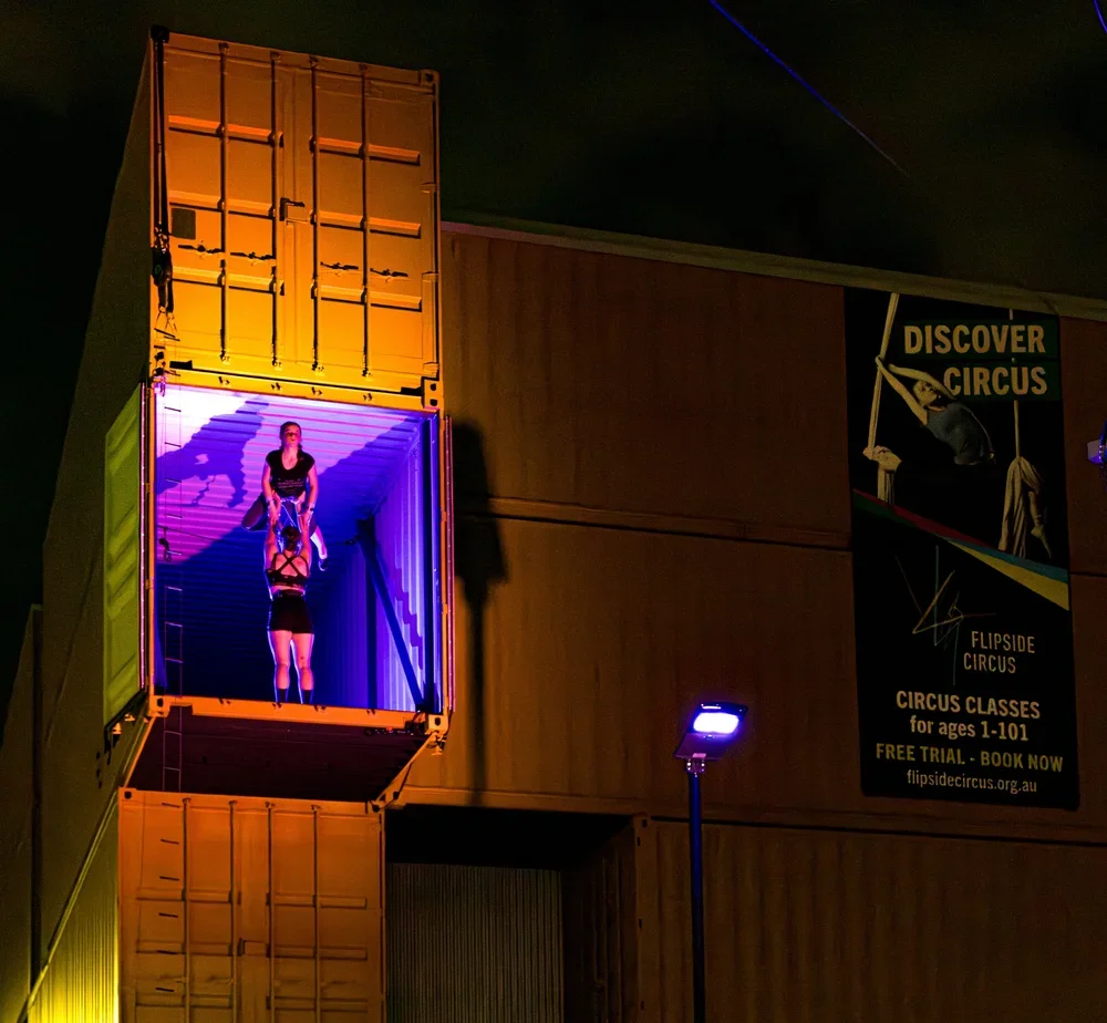A performer inside a shipping container illuminated with purple light, performing a balancing act on a suspended object at the Flipside Circus at night.