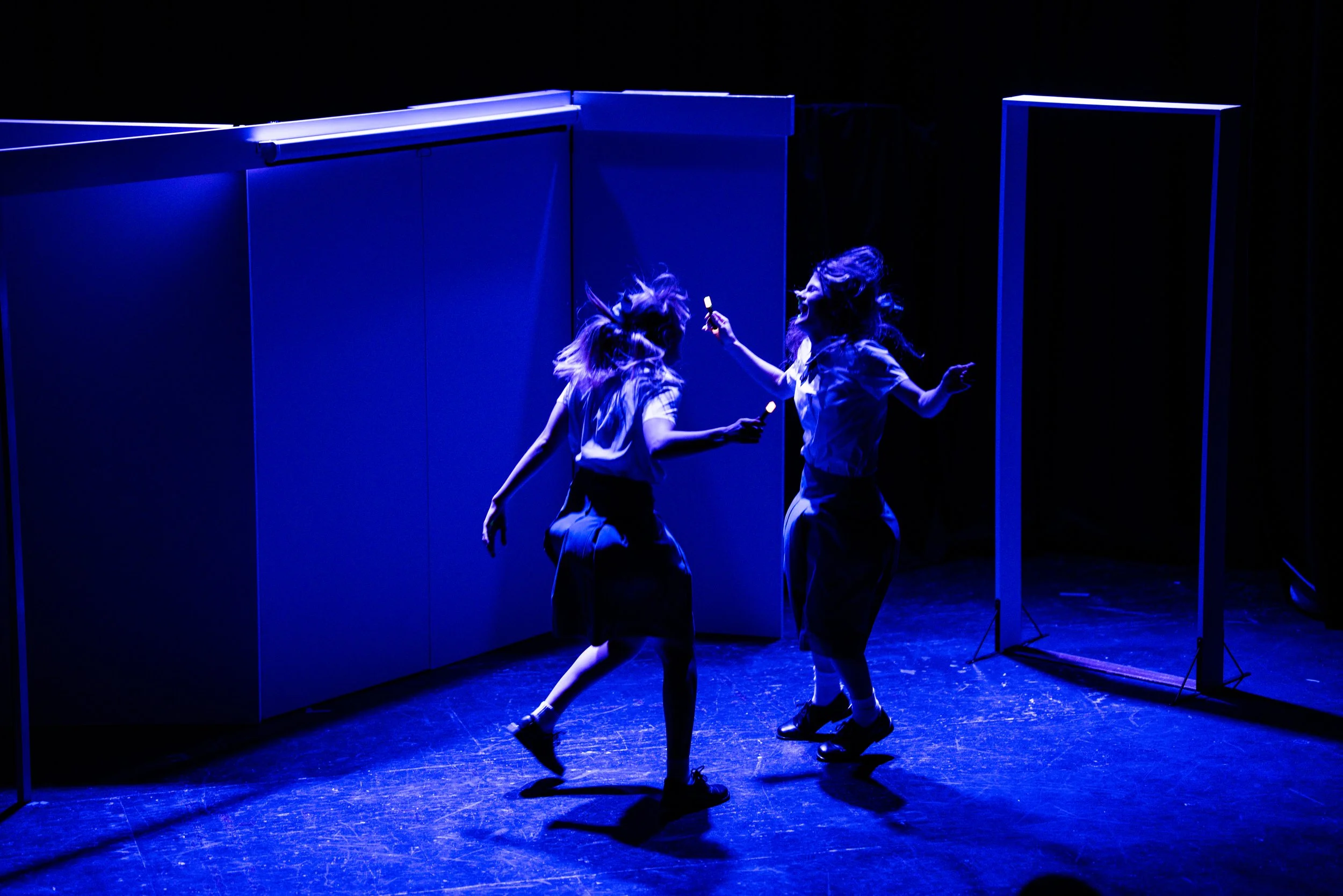 Two women dancing and laughing on a stage under blue lighting, holding markers. They are wearing similar school-like uniforms.