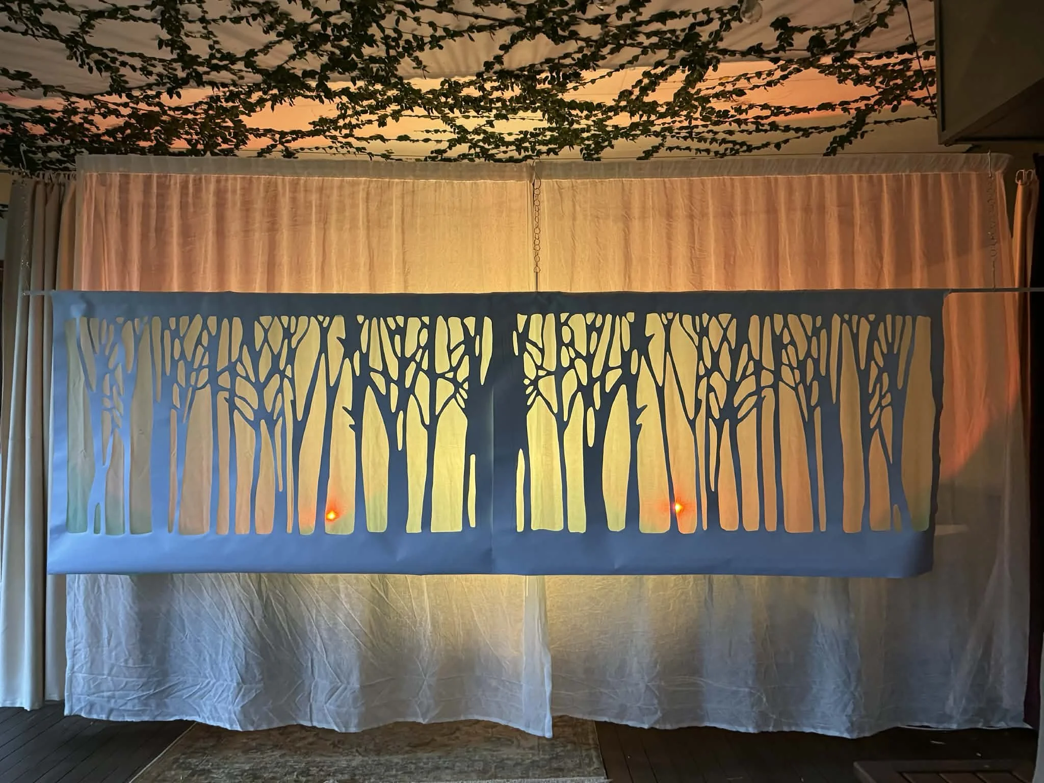 Decorative fabric panel with cut-out forest silhouette, illuminated by lights behind, hanging indoors with curtains and ceiling decorations in the background.