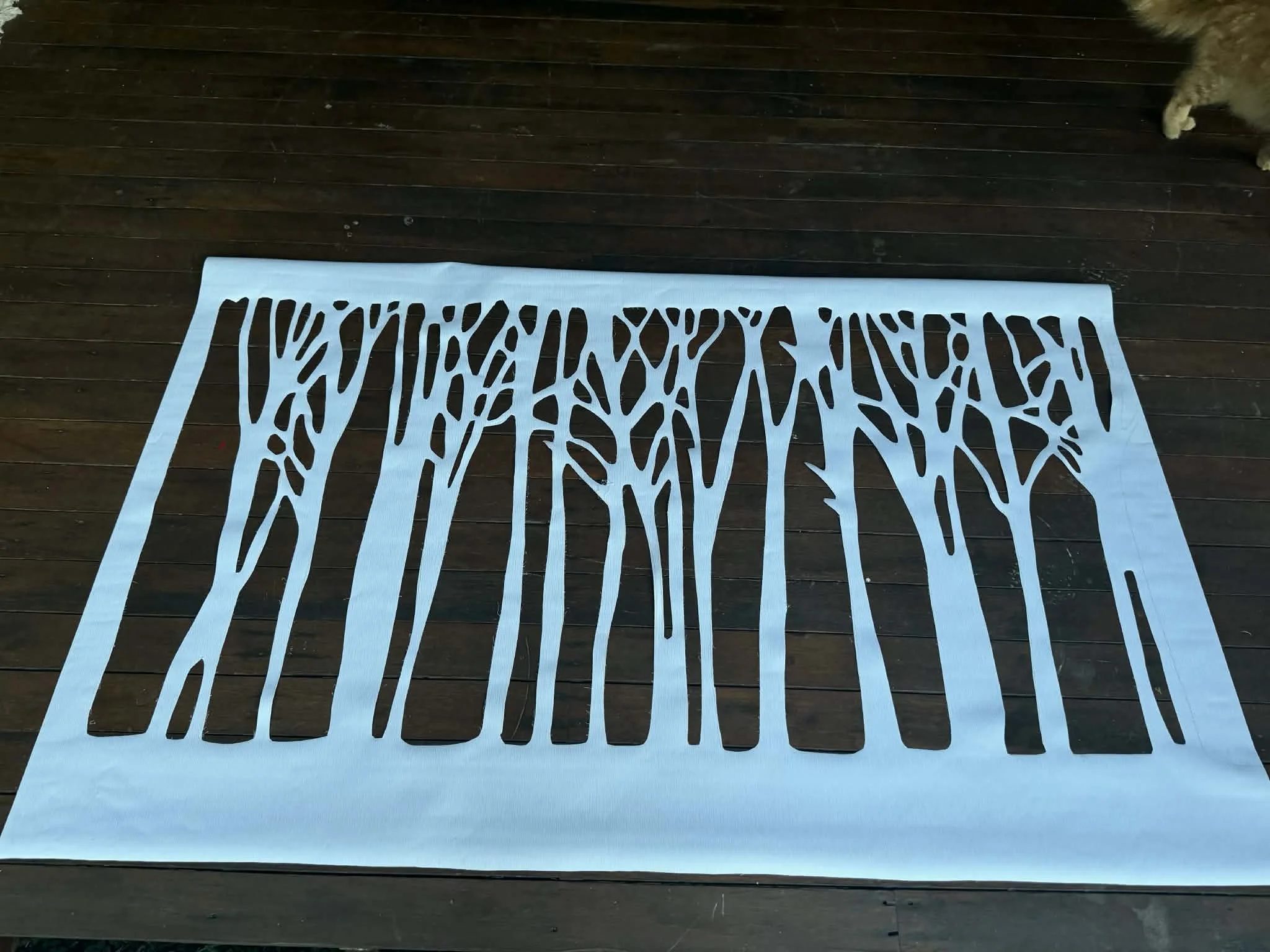 A white rectangular rug with black abstract tree branch patterns laid out on a wooden floor. Part of a dog is visible in the top right corner of the image.