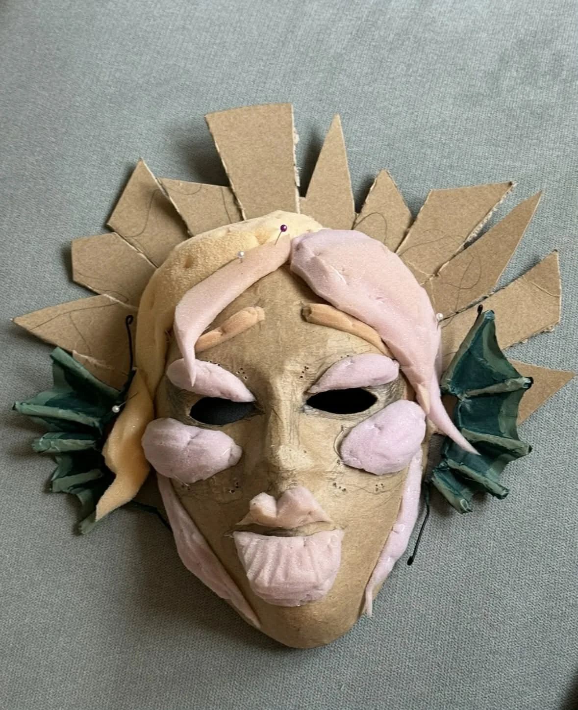 An artistic mask with a human face shape, decorated with paper and fabric elements, including pink and beige features, and surrounded by a cardboard sunburst or halo design.