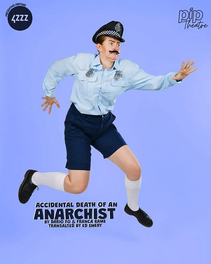 Person dressed as a police officer with a mustache, hat, and badge, jumping with arms extended, wearing shorts and white socks, against a solid purple background. Text on image reads 'Accidental Death of an Anarchist,' by Dario Fo & Franca Rame, tran