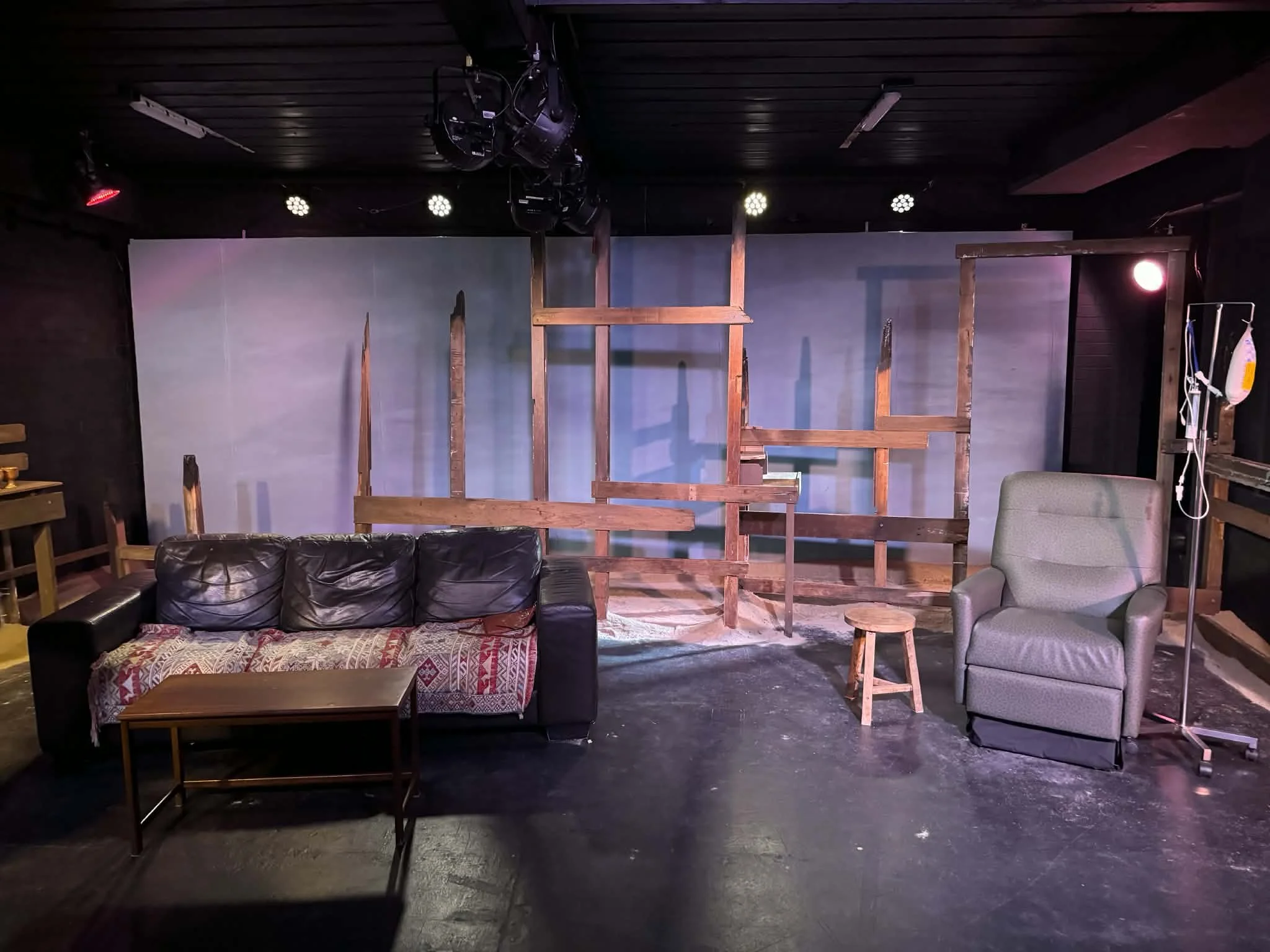 The stage set features a couch with a blanket, an armchair, a small stool, a wooden table, and a wooden scaffolding structure, with stage lights overhead.