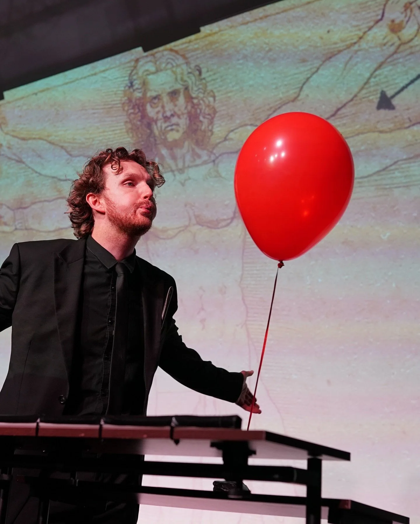 A man in a black suit holding a red balloon, with a projected image of Leonardo da Vinci's Vitruvian Man in the background.