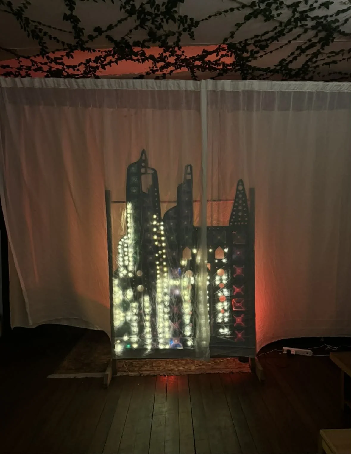 Art installation with illuminated cityscape silhouette featuring tall buildings and bridges, set against a warm background with hanging vines and a sheer curtain.