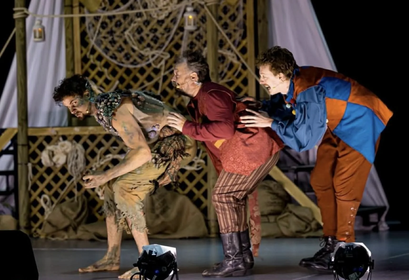 Three performers on stage in colorful costumes bent forward in a line, holding each other on the back, with a wooden lattice and hanging lights in background.