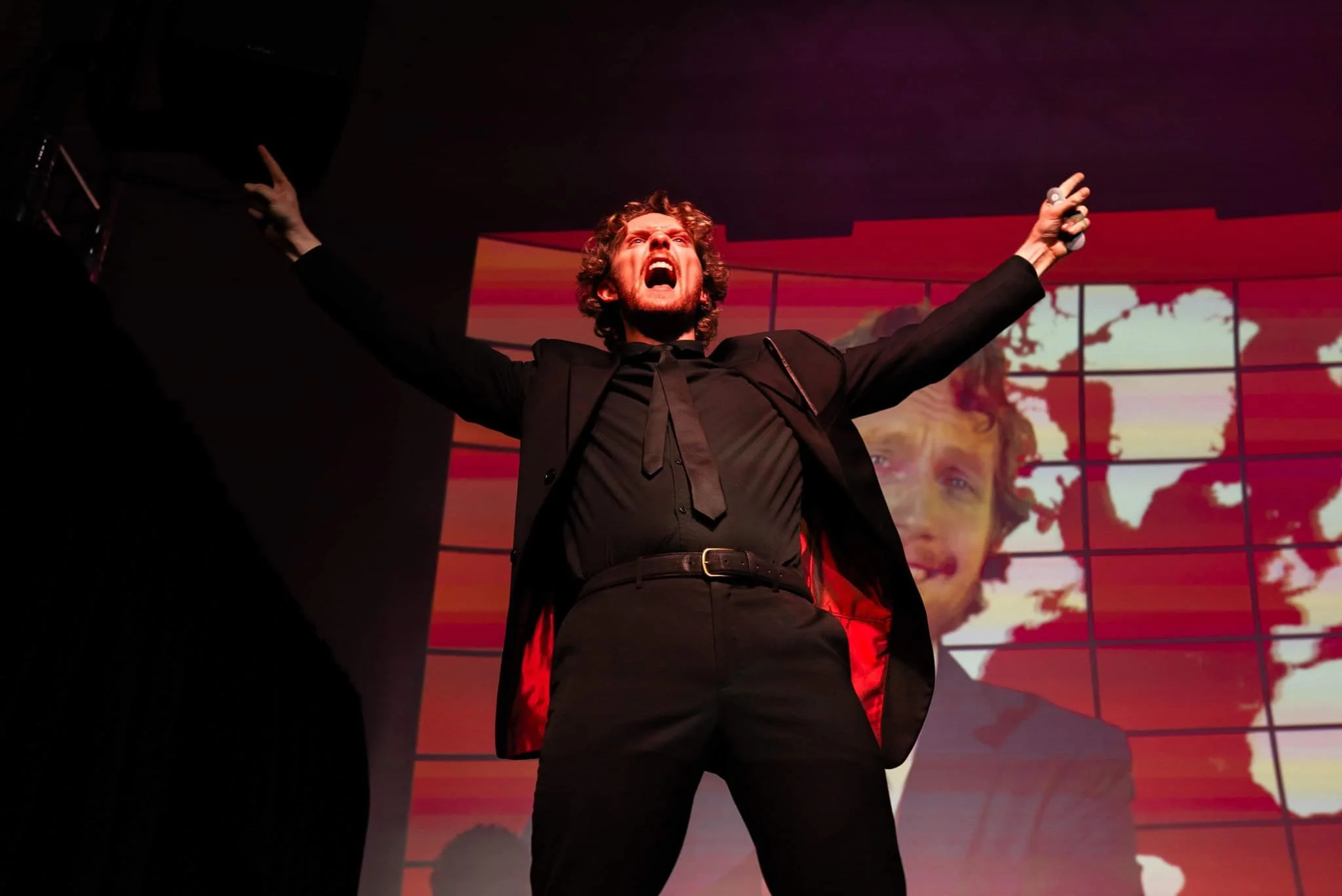 A man dressed in a black suit with a black shirt and a black tie, standing with arms outstretched and shouting, in front of a large screen displaying an animated world map with a smiling man's face in the background.