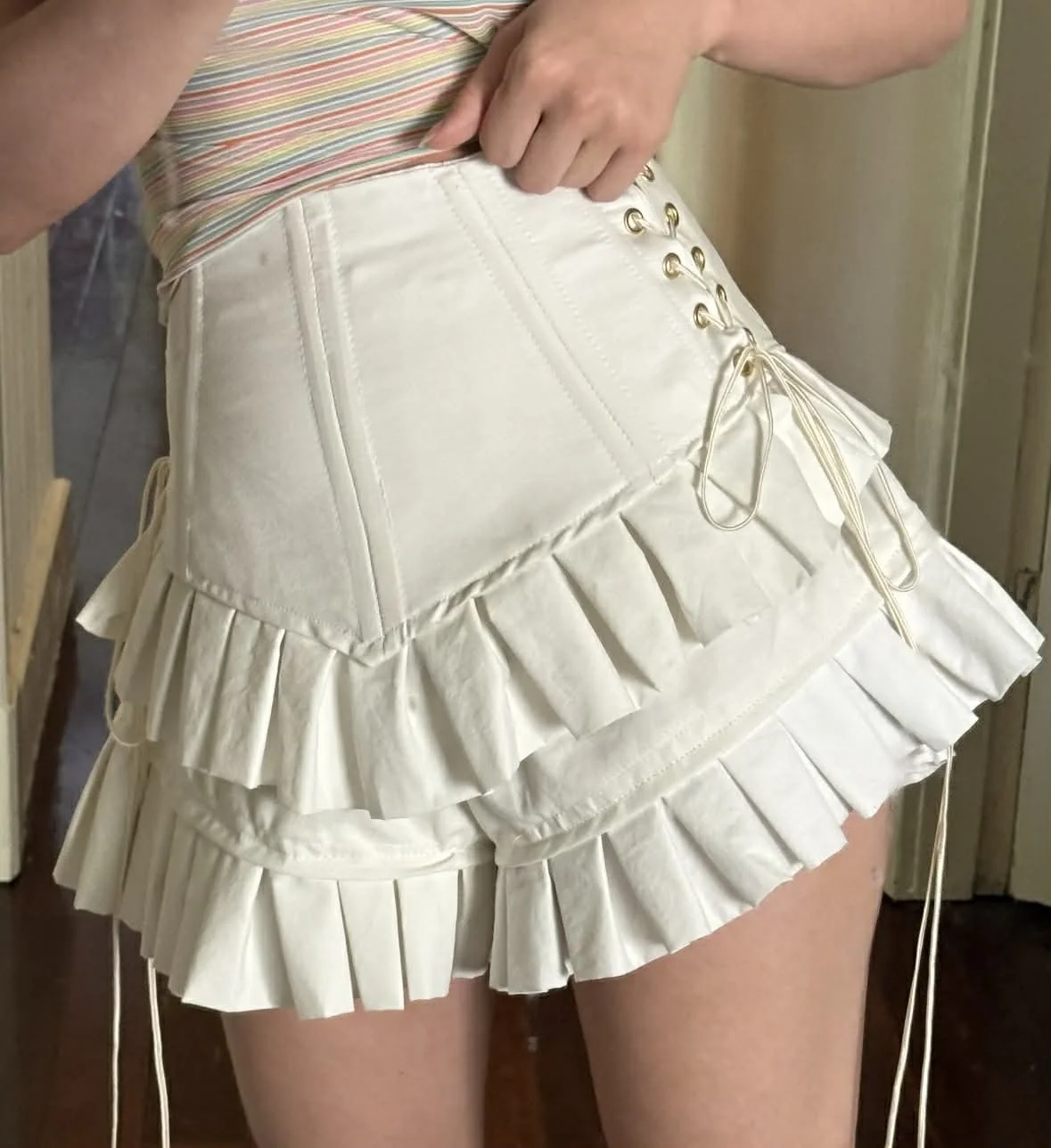 A person wearing a beige pleated skirt with lace-up details and a colorful striped top.