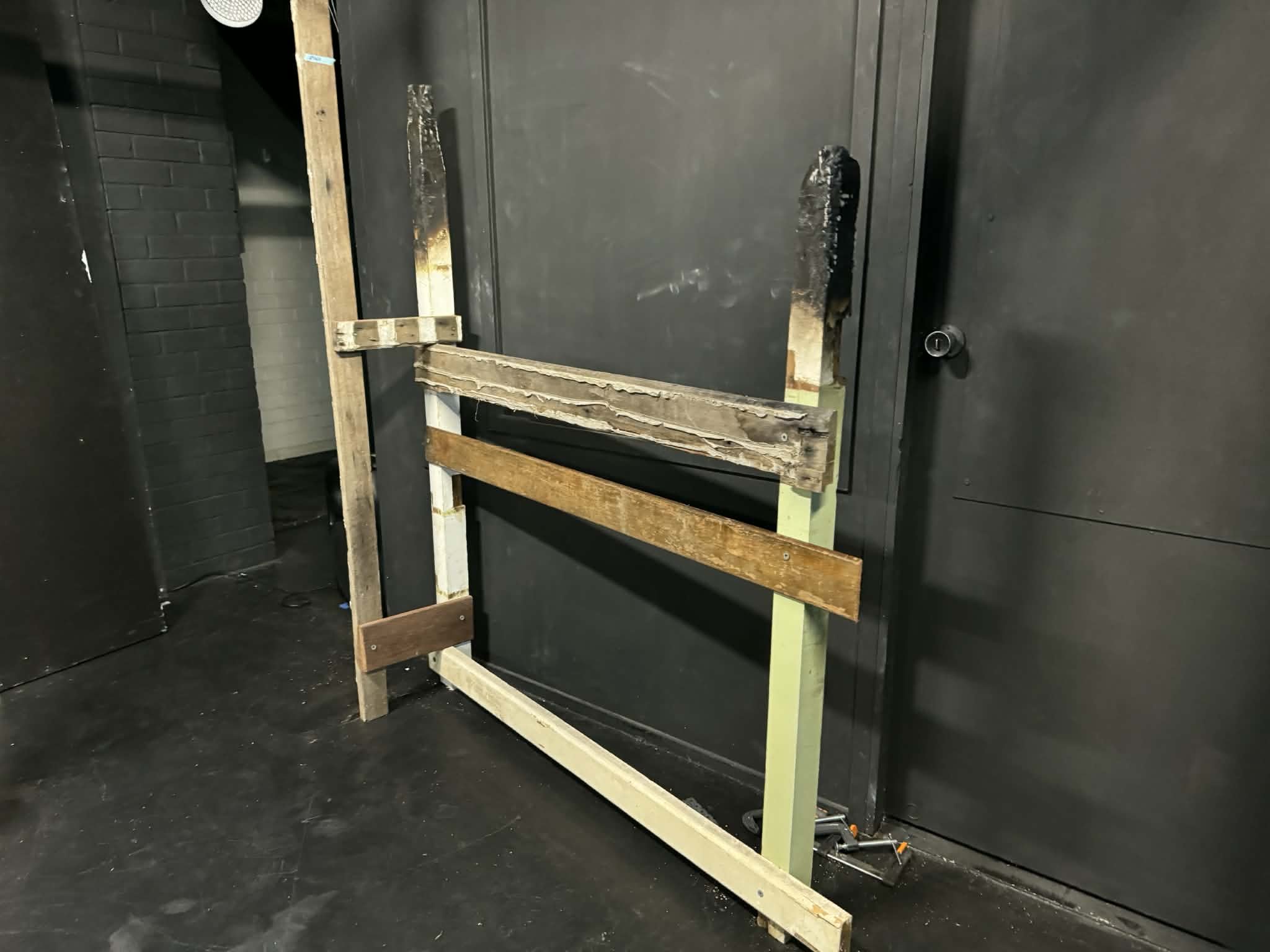 A partially assembled black metal door with damage and wooden supports holding it in place, situated in a dimly lit industrial or workshop setting.