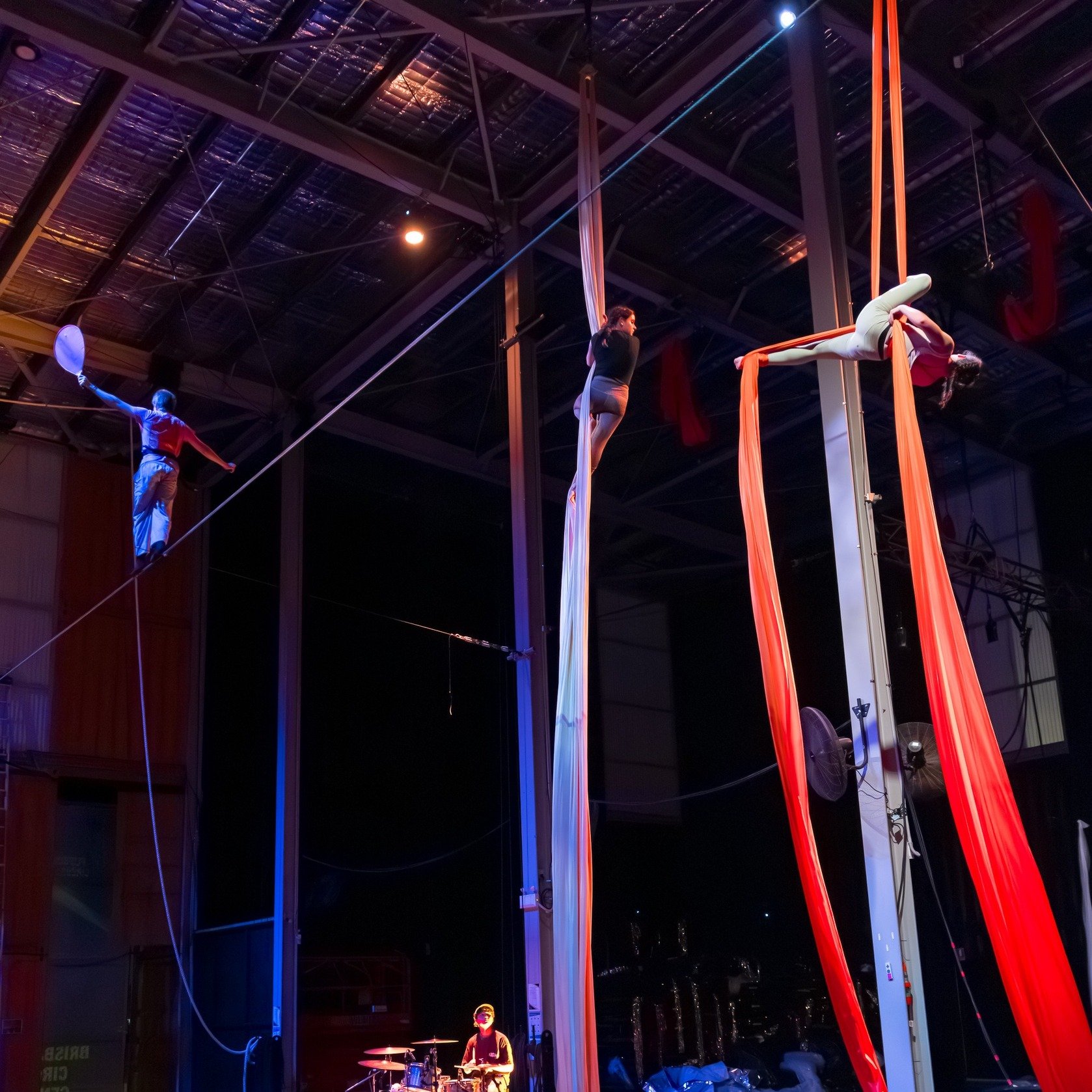 Two aerial performers practicing with long fabric silks in a large indoor space, with a circus performer on a tightrope and a drummer in the background.