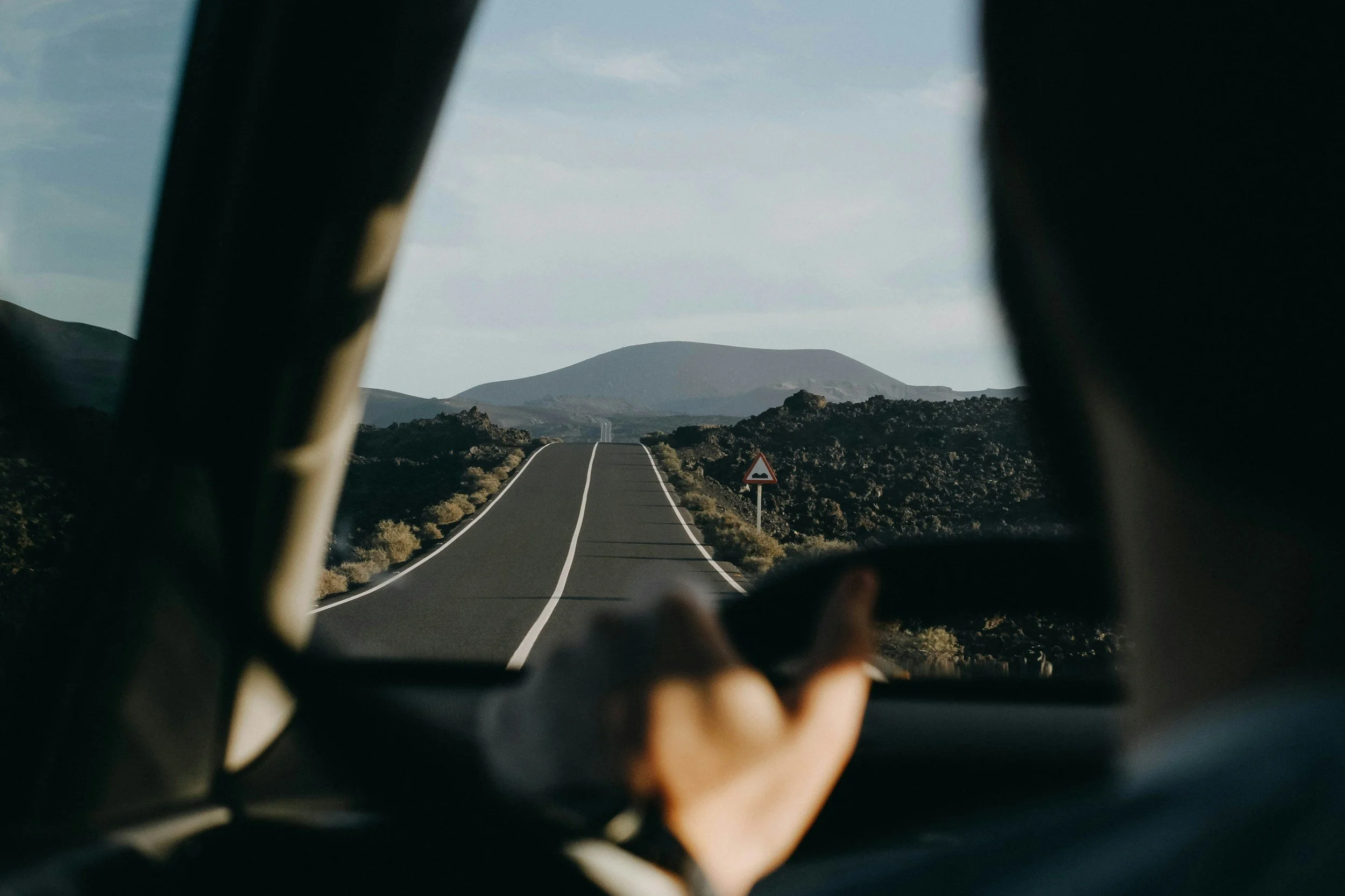 View from inside a car looking through the windshield at a winding road in a rural, mountainous landscape with a warning sign on the side of the road.