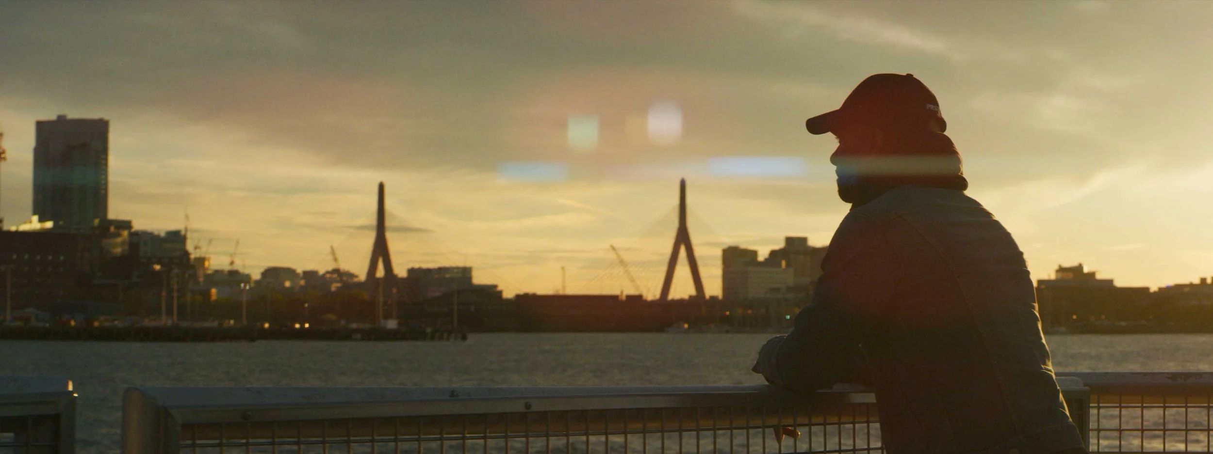 A commercial video image of man stands along the water at sunset looking at the boston skyline