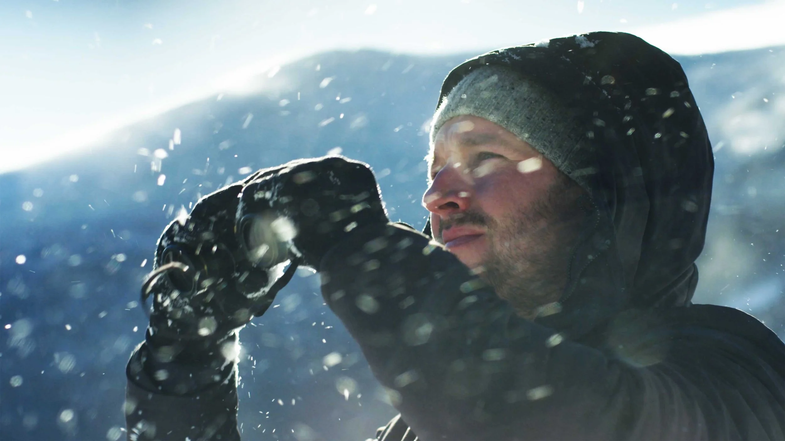 A man lifts binoculars as snow falls in the mountains