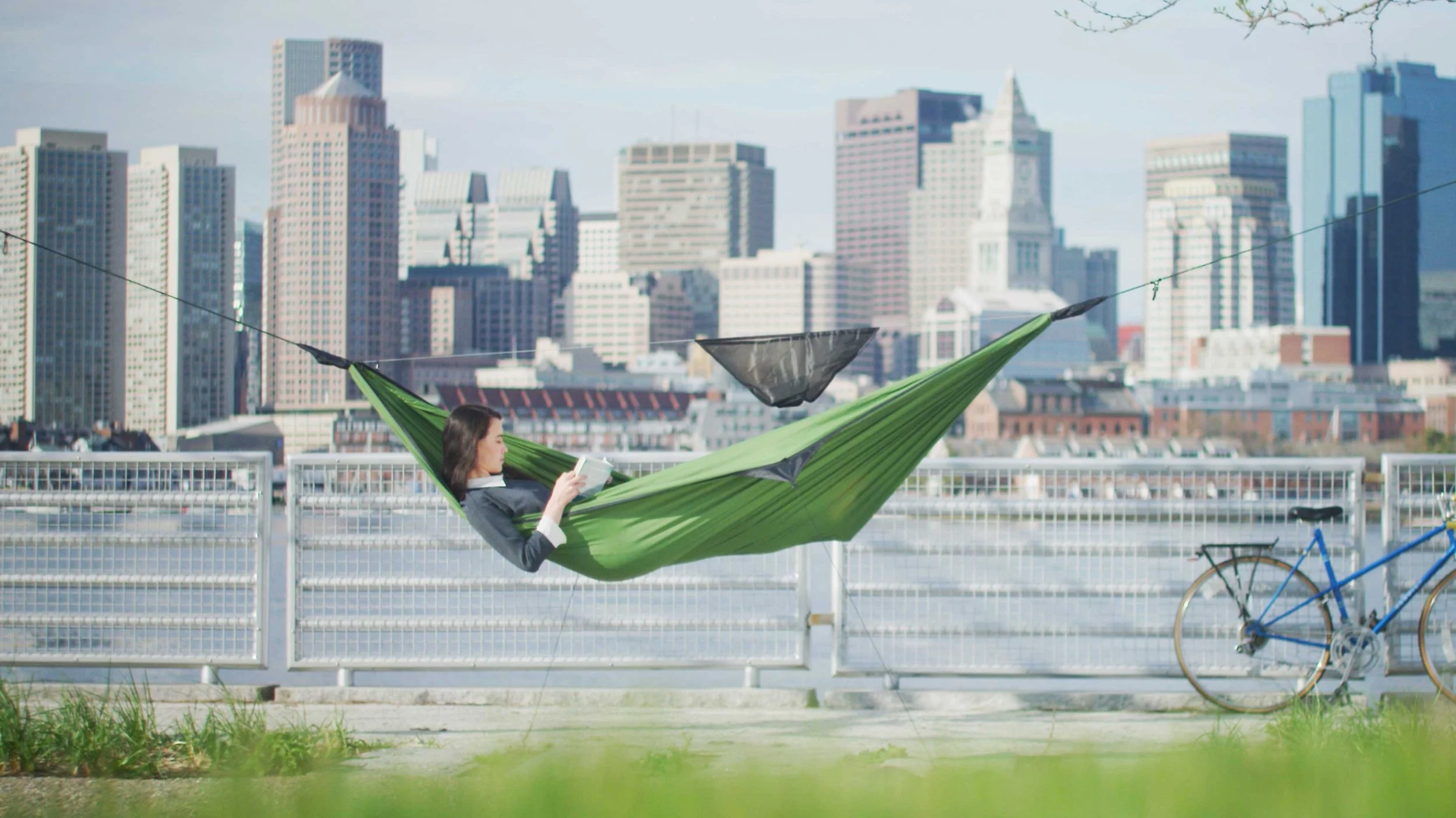 A woman sits in a Hennessy Hammock in front of the Boston skyline reading a book