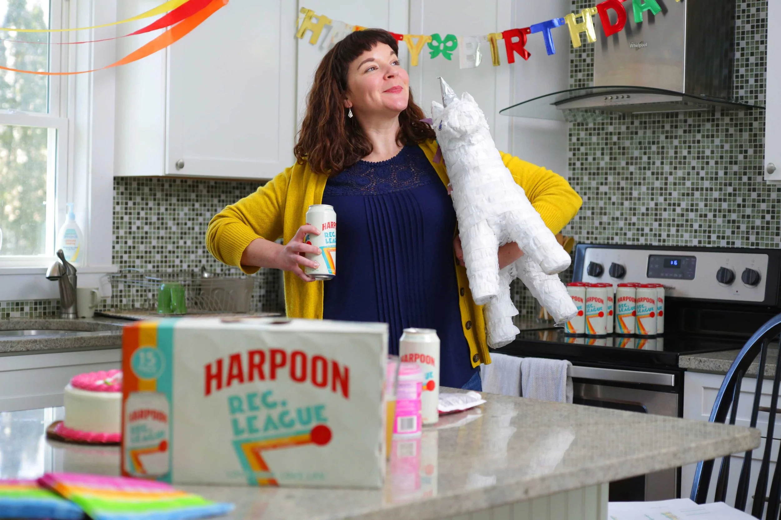 A woman stands in a kitchen proudly holding a unicorn piñata and a Harpoon Rec League IPA