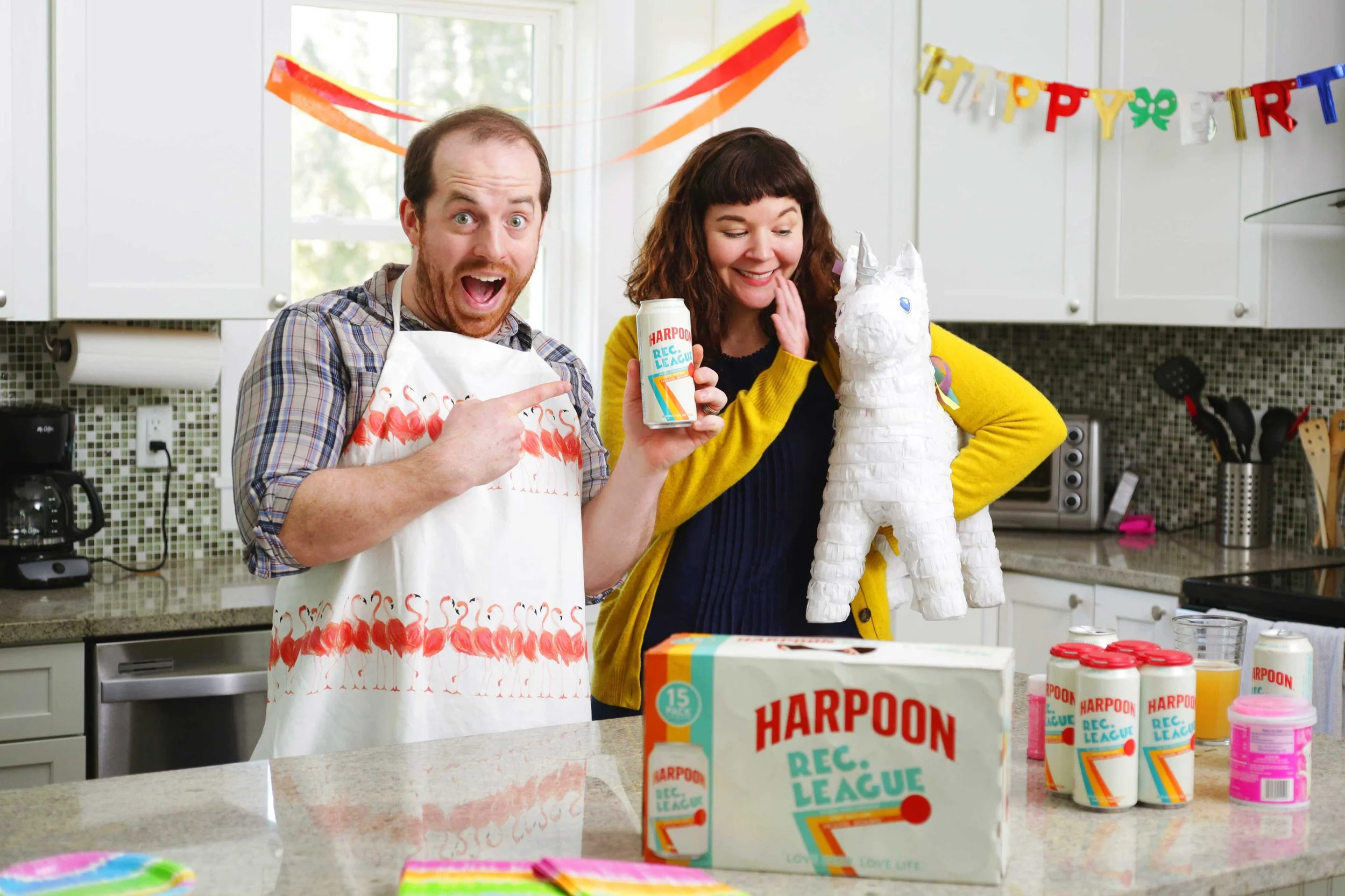 Man and woman stand in a kitchen, the man holds a Harpoon Lec League IPA and the woman holds a piñata