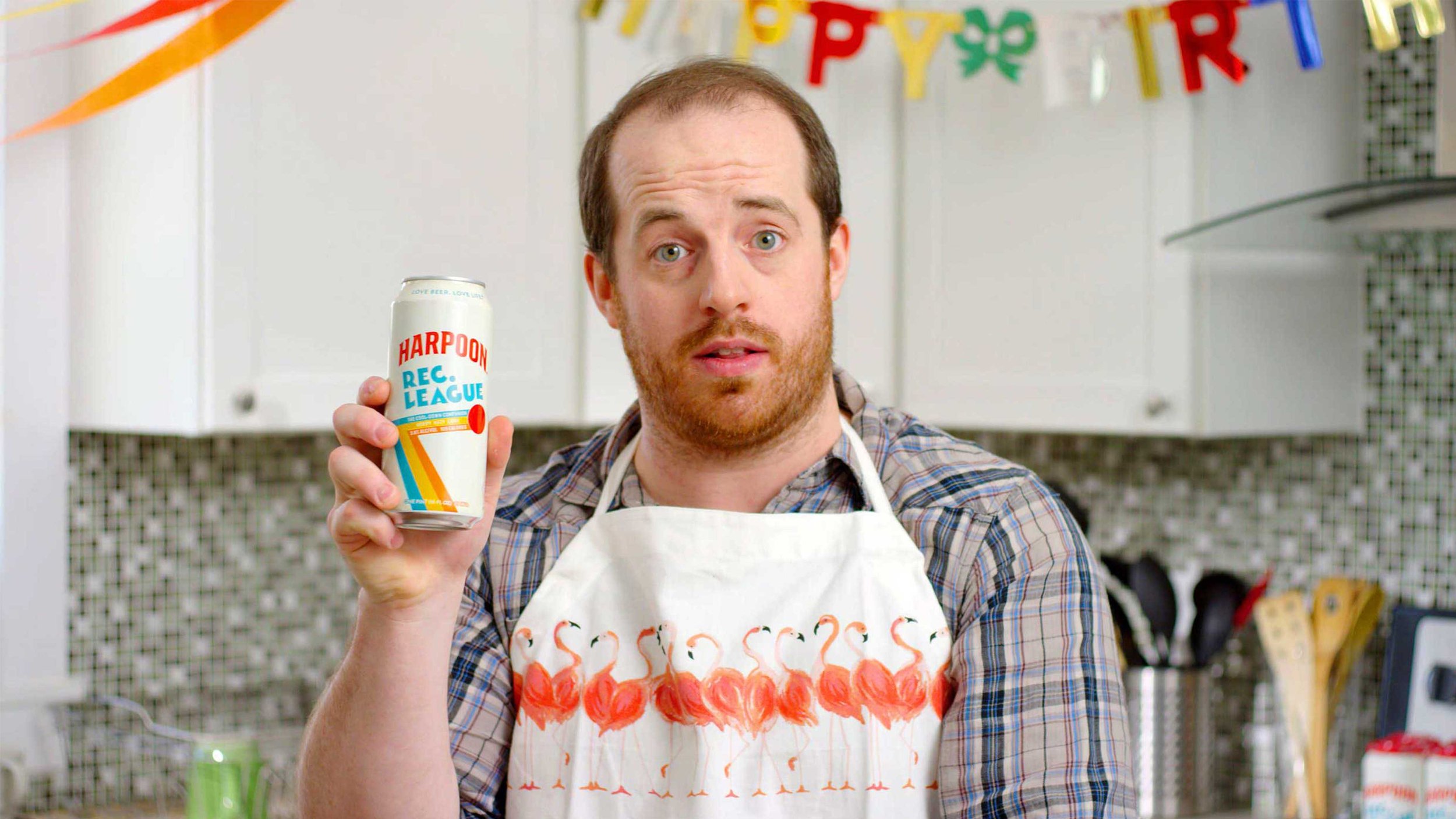 A man standing in a kitchen holding up a Harpoon Rec League IPA