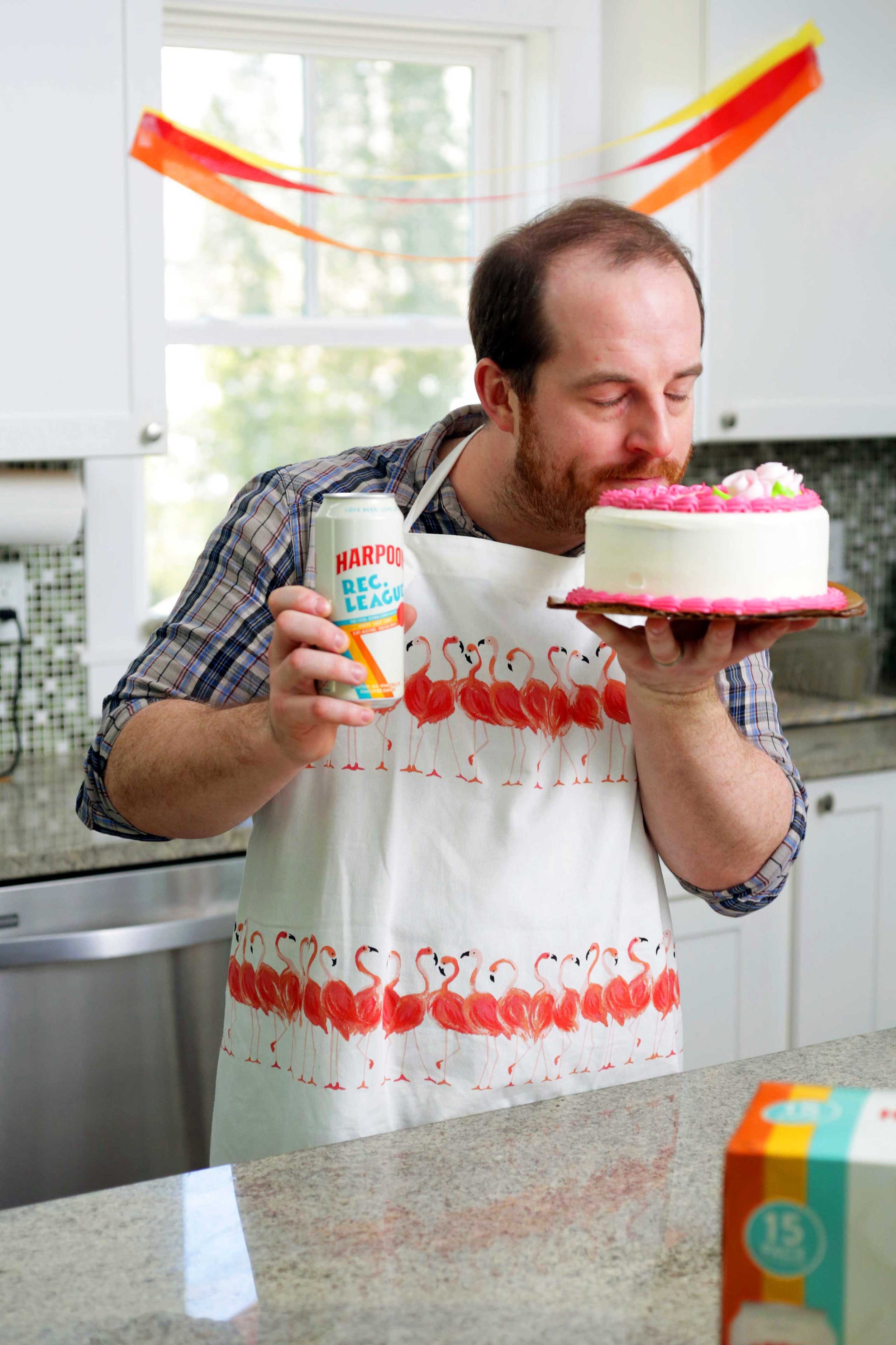 Man stands in a kitchen, holding a Harpoon Lec League IPA and sniffing a cake