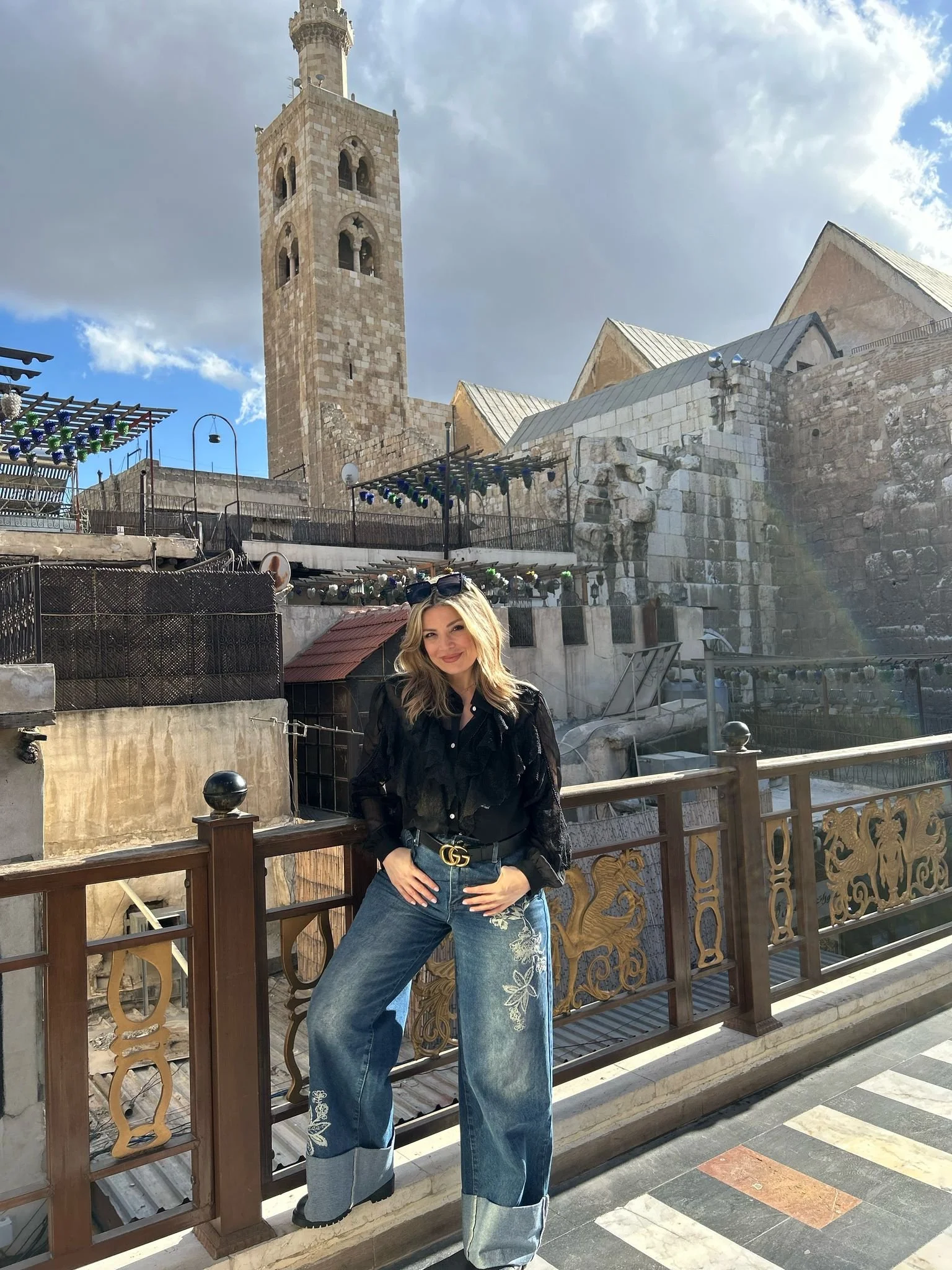 Woman standing on a bridge in front of a historic stone building with a tall clock tower, blue sky, and clouds visible.