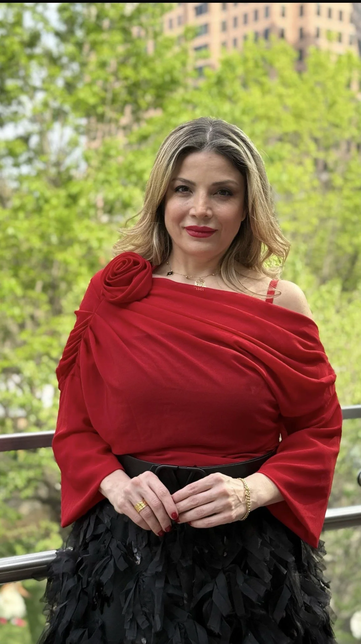 A woman with light brown wavy hair and red lipstick, wearing a red dress with a floral shoulder detail, black feathered skirt, and gold jewelry, standing outdoors with green trees and a building in the background.