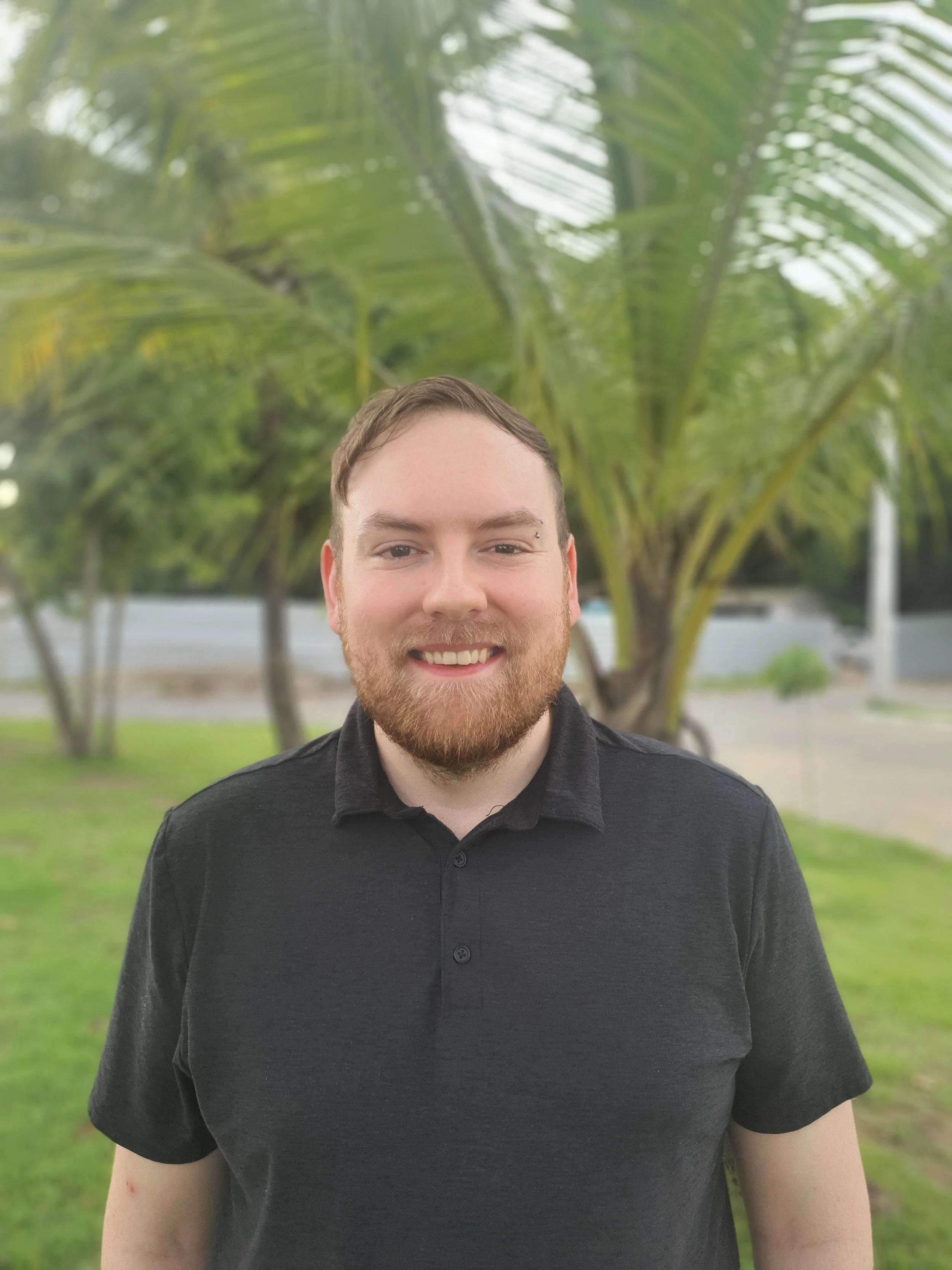 A smiling man with a beard and short hair standing outdoors in front of a large palm tree, wearing a black polo shirt.