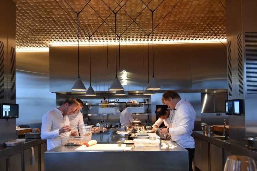 Chefs working in a professional kitchen with stainless steel appliances and hanging pendant lights.