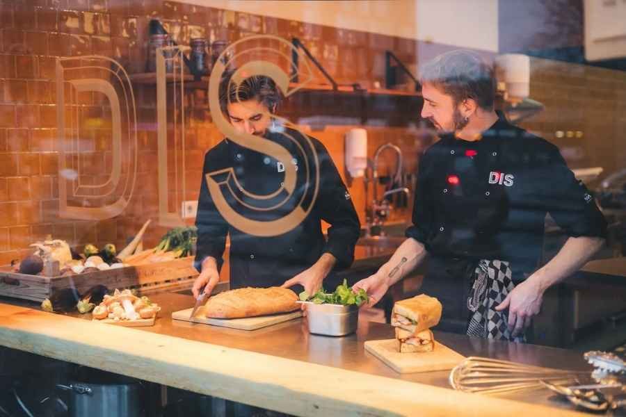 Two chefs in black uniforms preparing food in a kitchen, viewed through a glass door with a decorative logo. One is slicing bread, the other is holding a bowl of greens.
