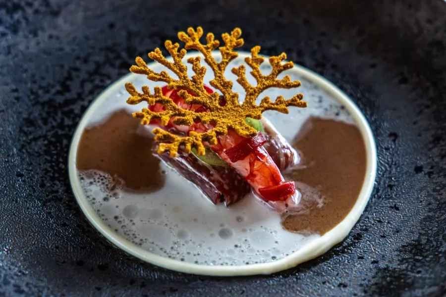 Close-up of gourmet beef dish in a black cast-iron skillet, featuring a piece of cooked beef topped with a decorative yellow garnish and surrounded by a creamy sauce with foam.