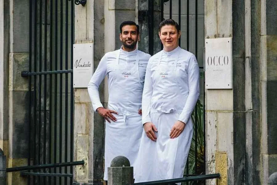 Two male chefs standing outdoors in front of a stone building with black iron gates, dressed in white chef uniforms and aprons, smiling at the camera.