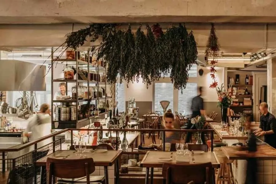 Interior of a cozy restaurant with hanging dried herbs and flowers, wooden tables set with glasses and cutlery, and people dining and working in the background.