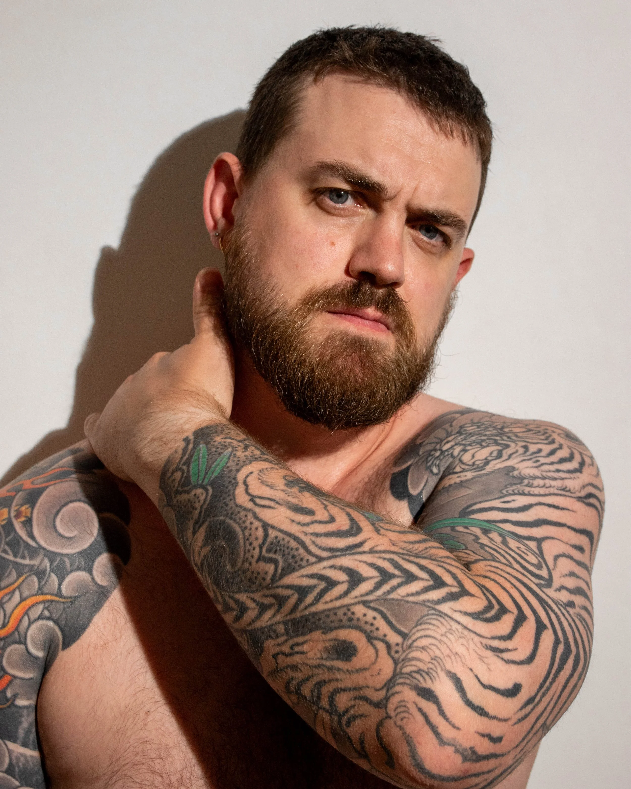 A shirtless man with a beard and tattoos on his arms, looking serious, with his hand on the back of his neck, standing against a plain white background.